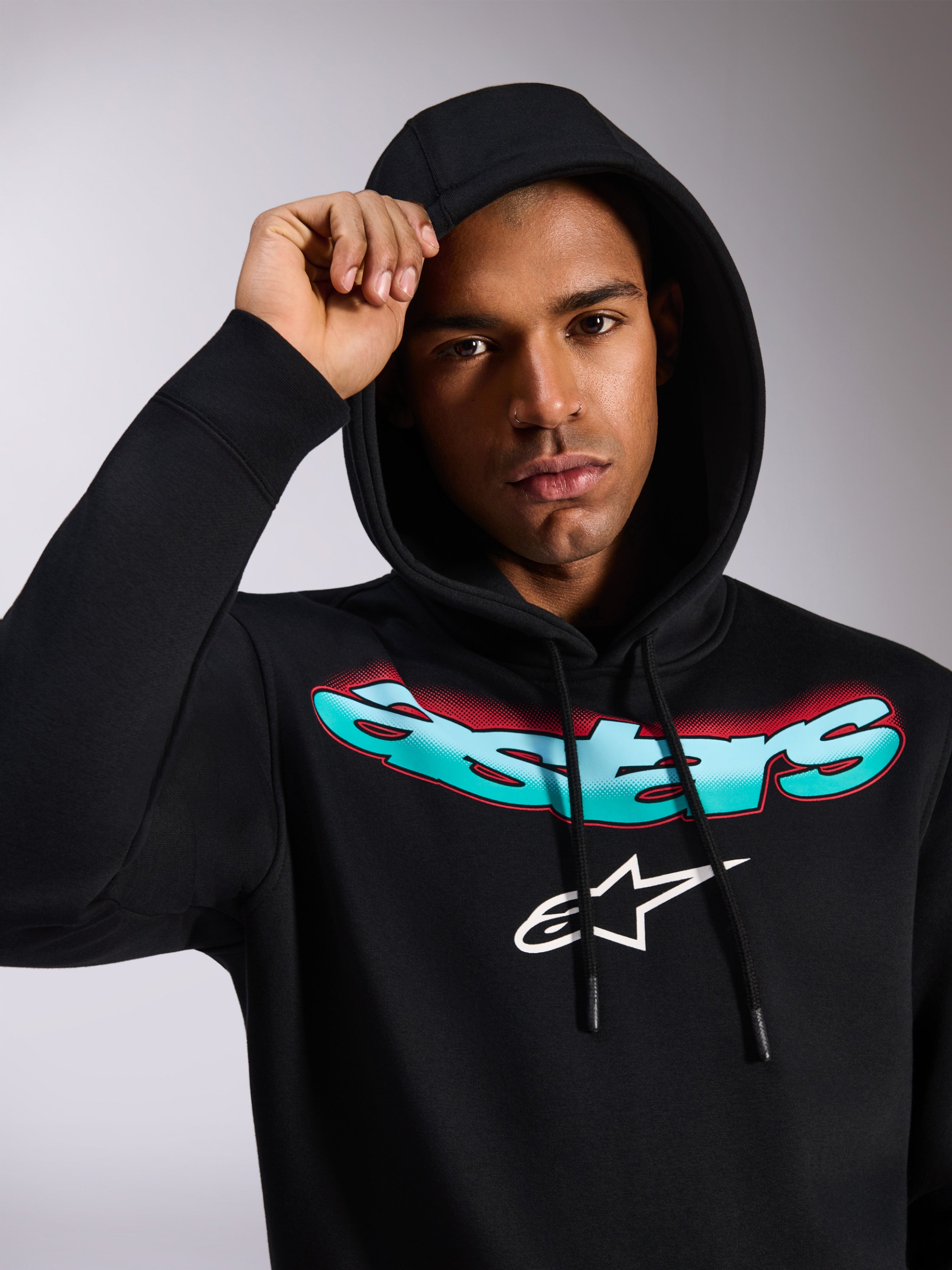 Alpinestars Ellipsoid Hoodie, felpa in pile nera a pullover, con scritta Astars stilizzata e sfumata verde acqua e rosso e logo bianco sul petto, indossata da un modello con cappuccio alzato, streetwear casual ispirato al motorsport