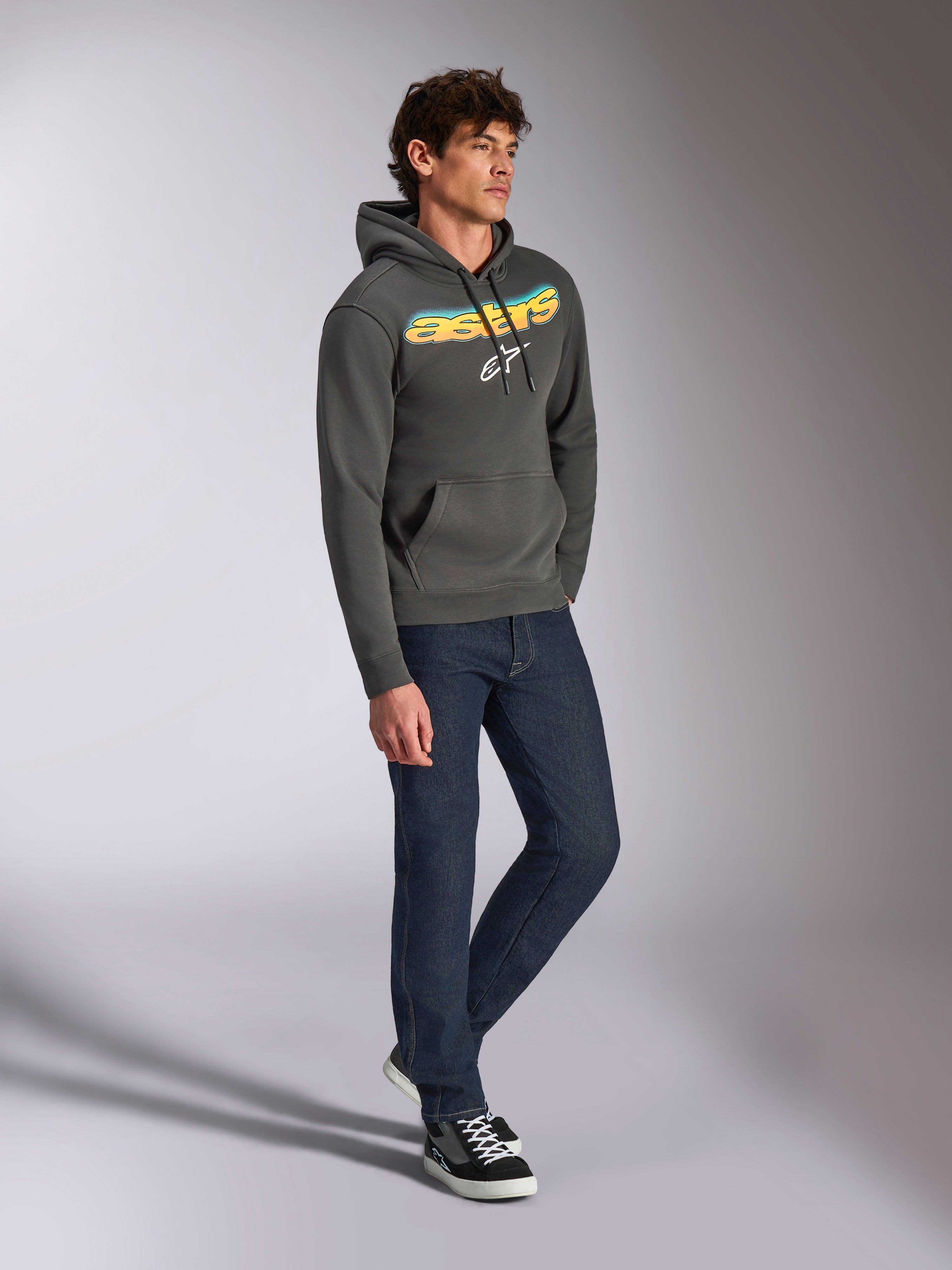 Felpa con cappuccio Alpinestars Ellipsoid, pullover in pile grigio canna di fucile, scritta frontale curva Astars con gradiente blu e giallo e logo bianco, abbinata a jeans scuri e sneakers nere, abbigliamento casual streetwear.