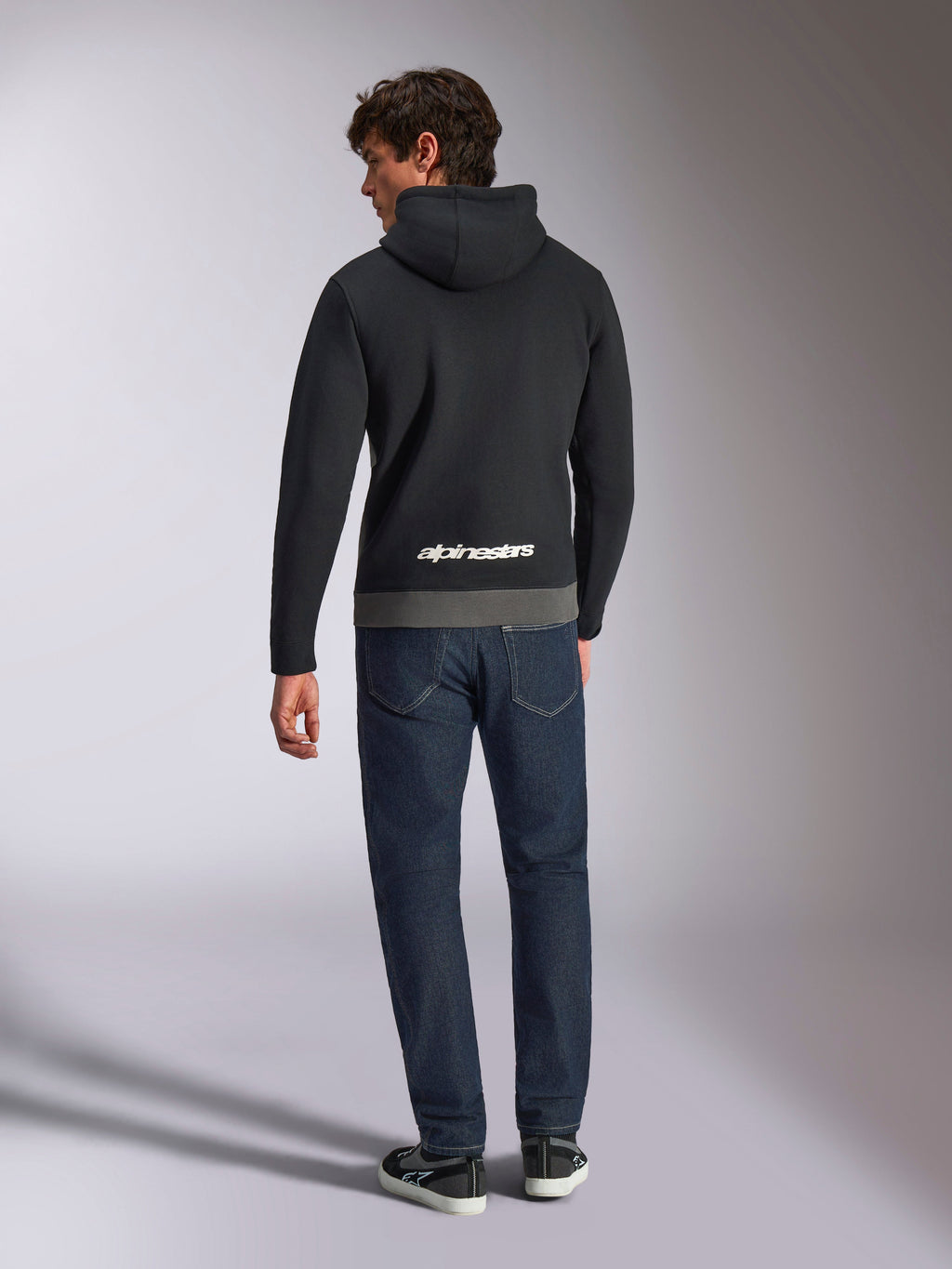 Felpa con cappuccio Alpinestars Parallel, vista posteriore di un uomo che indossa un pullover in pile a blocchi di colore nero e gunmetal, logo Alpinestars bianco sulla parte inferiore della schiena, abbinata a jeans blu scuro, streetwear casual ispirato al motorsport