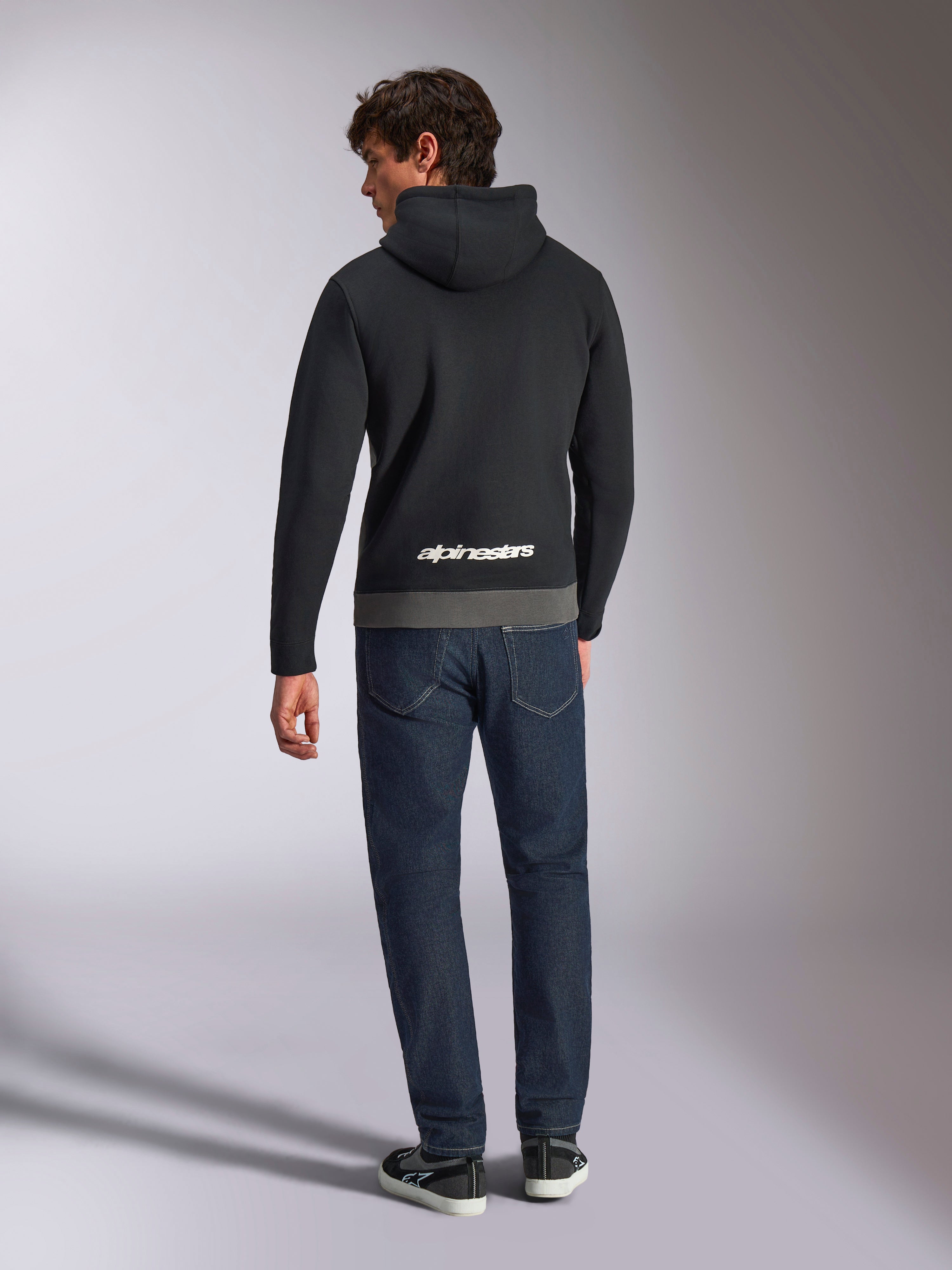 Felpa con cappuccio Alpinestars Parallel, vista posteriore di un uomo che indossa un pullover in pile a blocchi di colore nero e gunmetal, logo Alpinestars bianco sulla parte inferiore della schiena, abbinata a jeans blu scuro, streetwear casual ispirato al motorsport