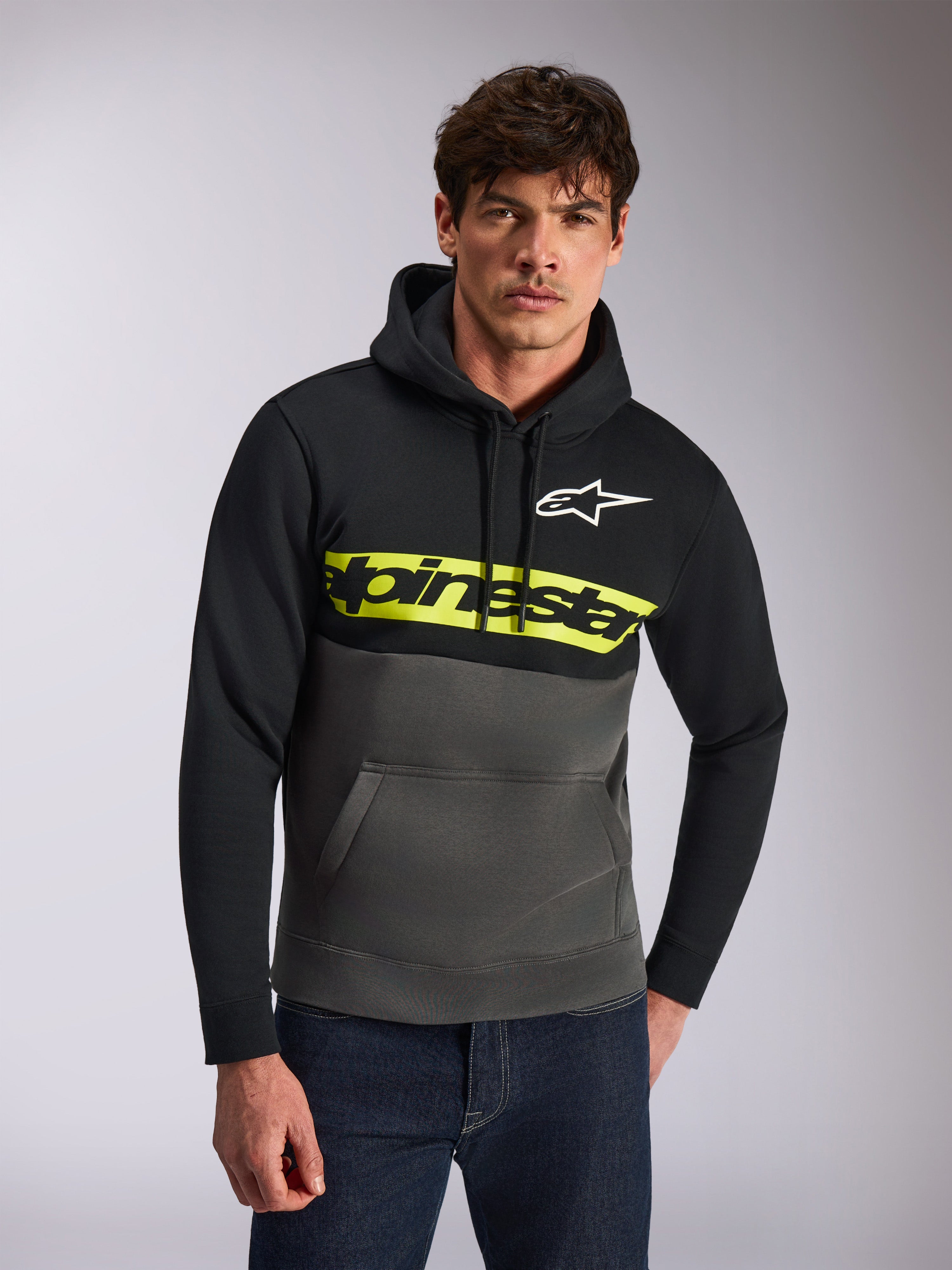 Alpinestars Parallel Hoodie, felpa con cappuccio casual, a blocchi di colore nero e grigio canna di fucile, striscia gialla brillante sul petto con logo Alpinestars, logo Astars bianco, indossata da un modello con jeans blu, design moderno ispirato al motorsport