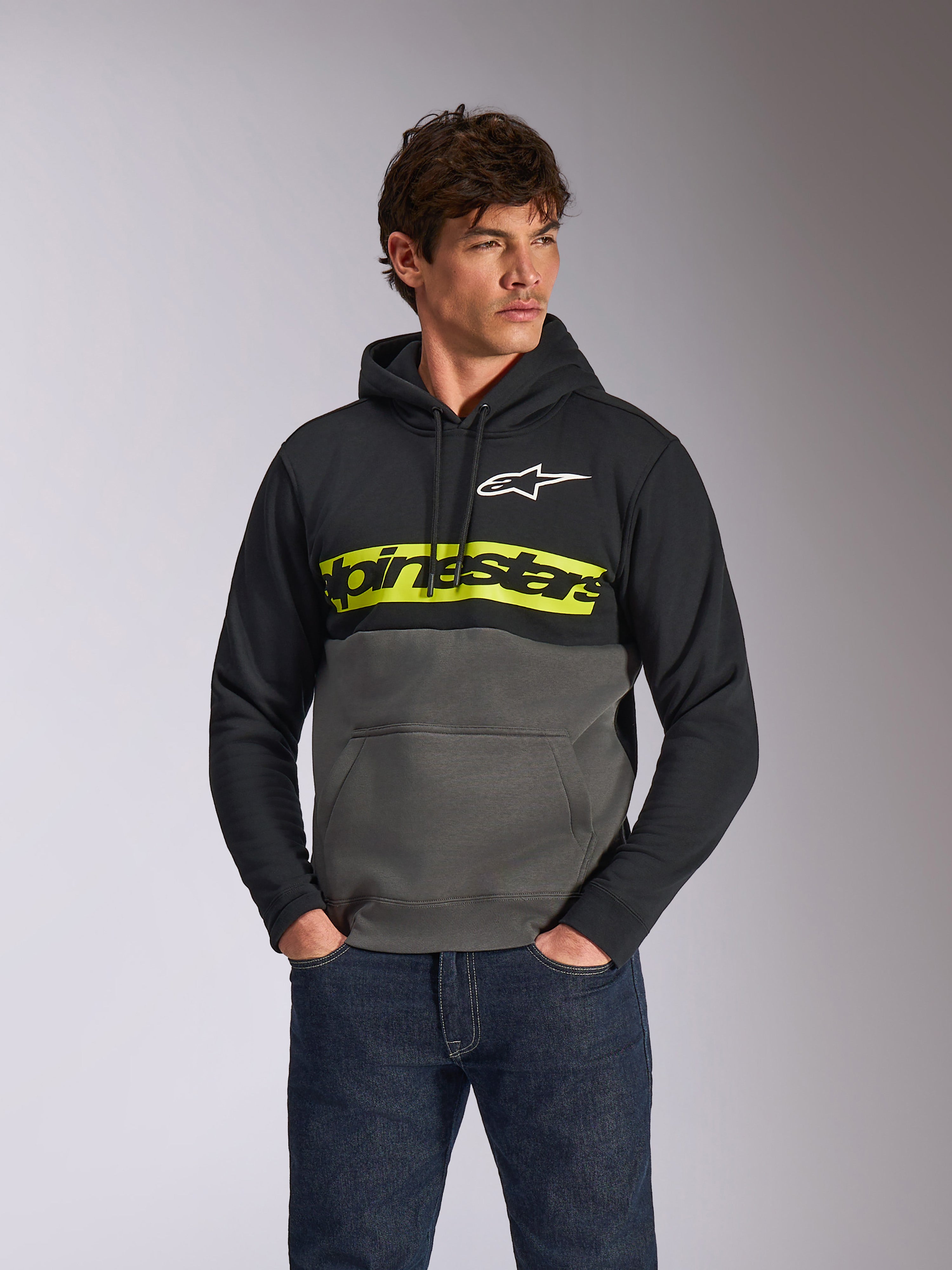 Alpinestars Parallel Hoodie, felpa pullover casual a blocchi di colore, nero e grigio canna di fucile con una barra gialla vivace sul petto, logo Alpinestars e logo a stella bianco, indossata da un modello con jeans blu scuro