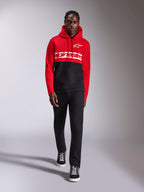 Felpa con cappuccio Alpinestars Parallel, felpa in pile a blocchi di colore, rosso e nero, con logo Alpinestars bianco sul petto e logo Astars sul petto a sinistra, indossata da un modello con pantaloni neri e sneakers alte, abbigliamento casual motorsport streetwear