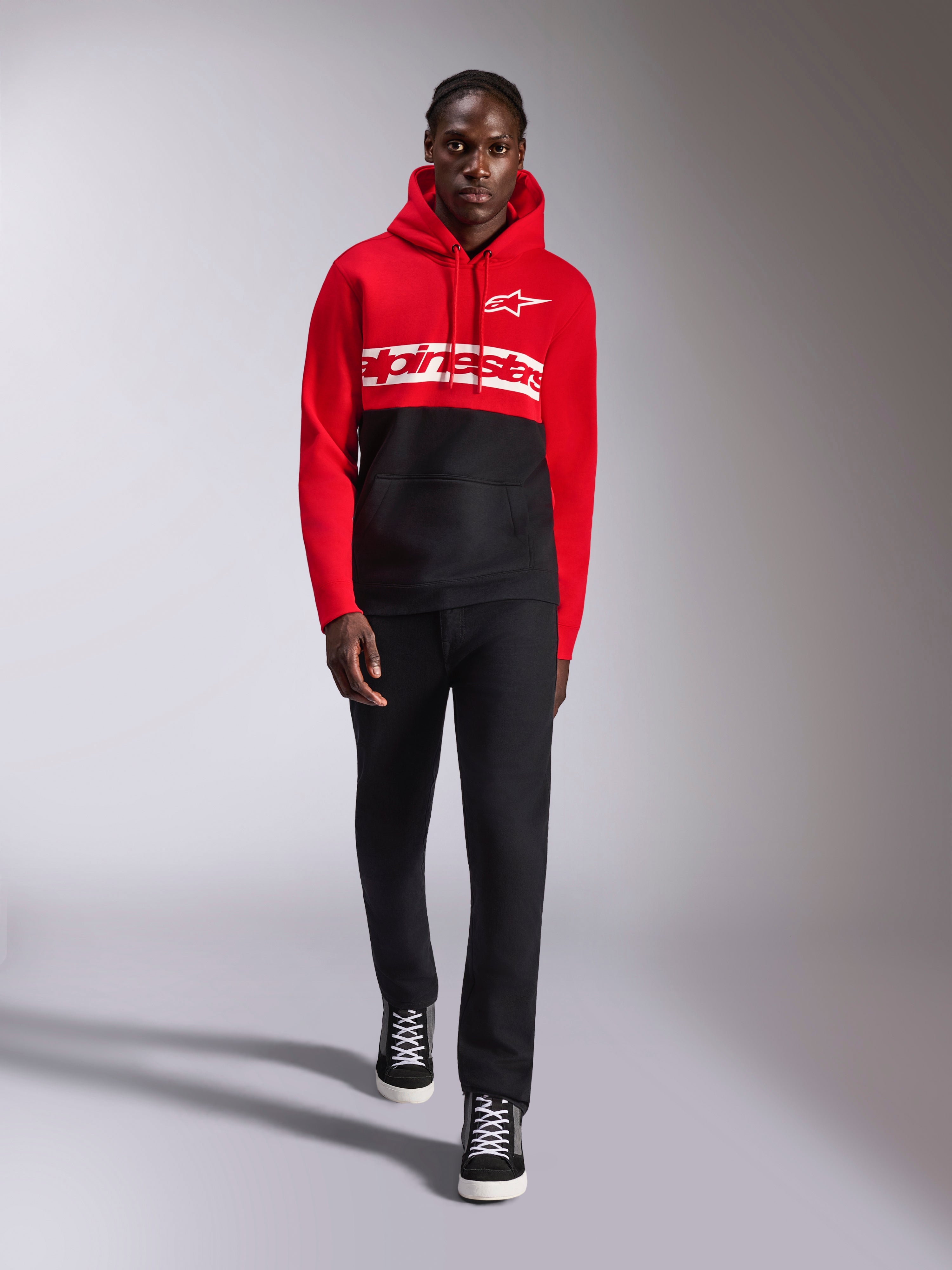 Felpa con cappuccio Alpinestars Parallel, felpa in pile a blocchi di colore, rosso e nero, con logo Alpinestars bianco sul petto e logo Astars sul petto a sinistra, indossata da un modello con pantaloni neri e sneakers alte, abbigliamento casual motorsport streetwear