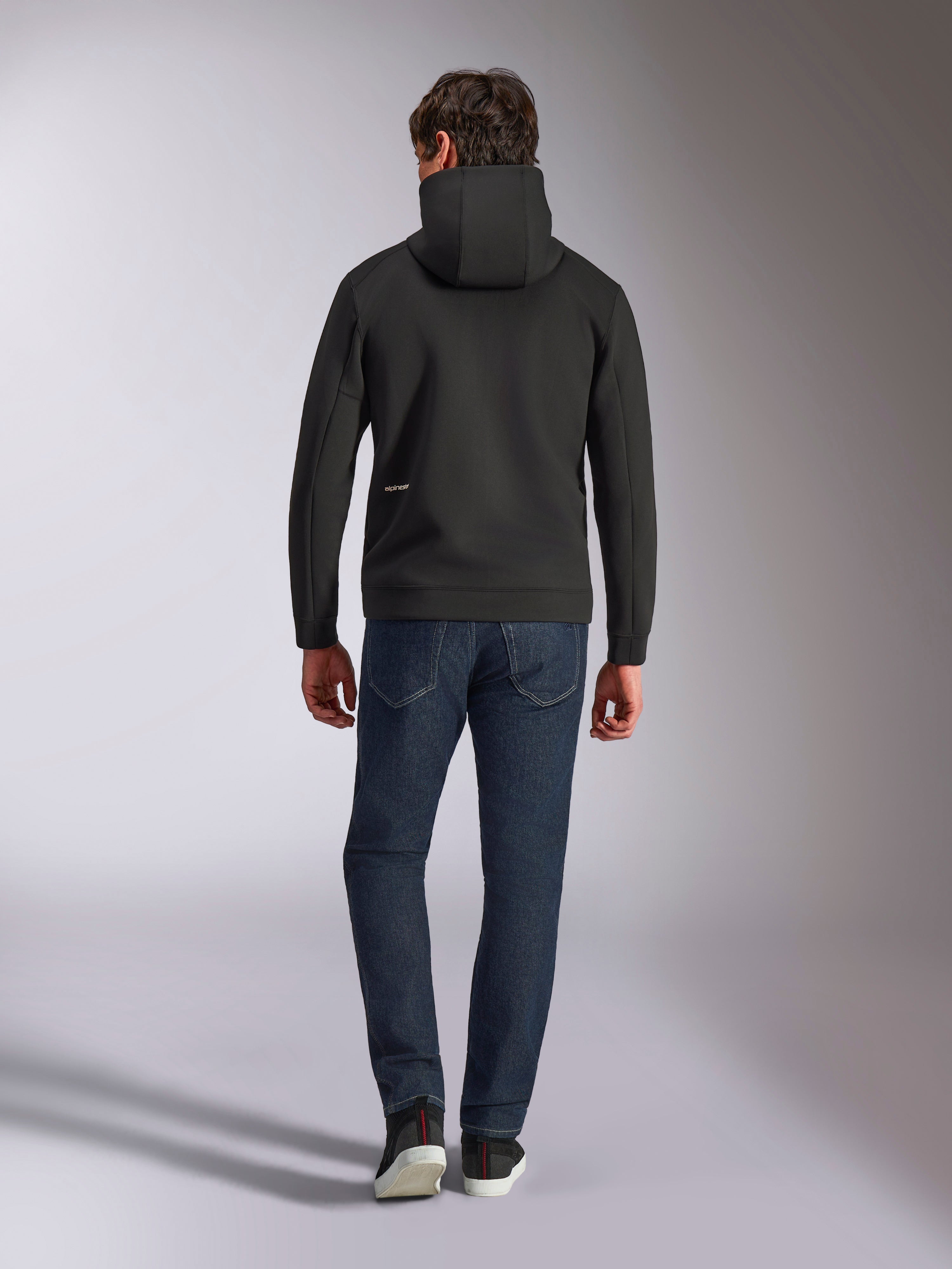 Alpinestars Cypher Hoodie, pile double-face nero, vista posteriore con piccolo logo Alpinestars bianco sul fianco sinistro, cappuccio a collo alto, abbinata a jeans blu scuro, streetwear casual ispirato al motorsport