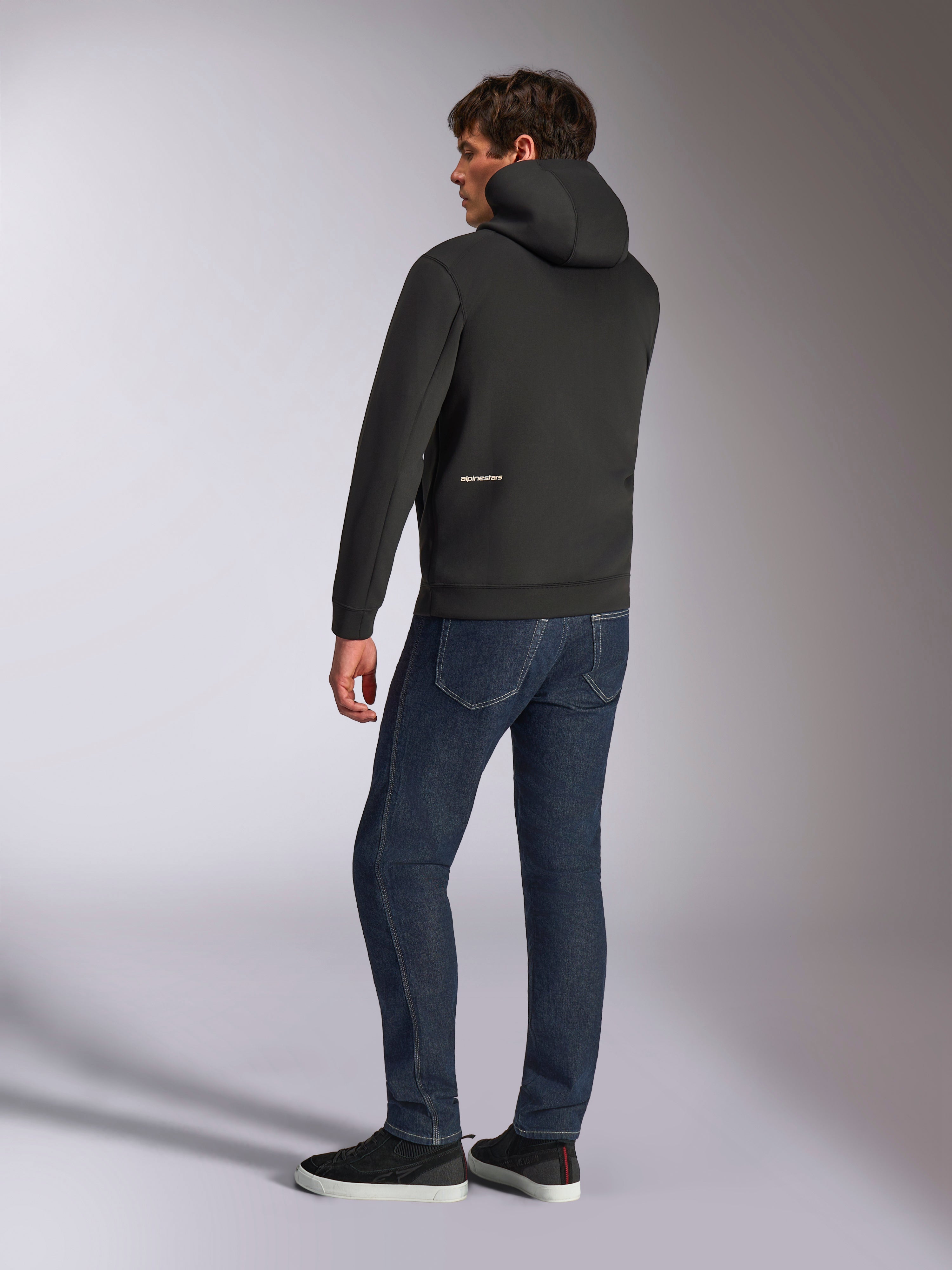 Alpinestars Cypher Hoodie, felpa casual nera con zip, vista posteriore con cappuccio a collo alto e logo Alpinestars bianco sulla parte bassa della schiena, indossata con jeans blu scuro e sneakers nere, design moderno ispirato al motorsport