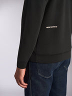 Felpa con cappuccio Alpinestars Cypher, vista ravvicinata della parte bassa della schiena e della manica, pile double-face nero con logo Alpinestars bianco, indossata con jeans blu, streetwear casual motorsport