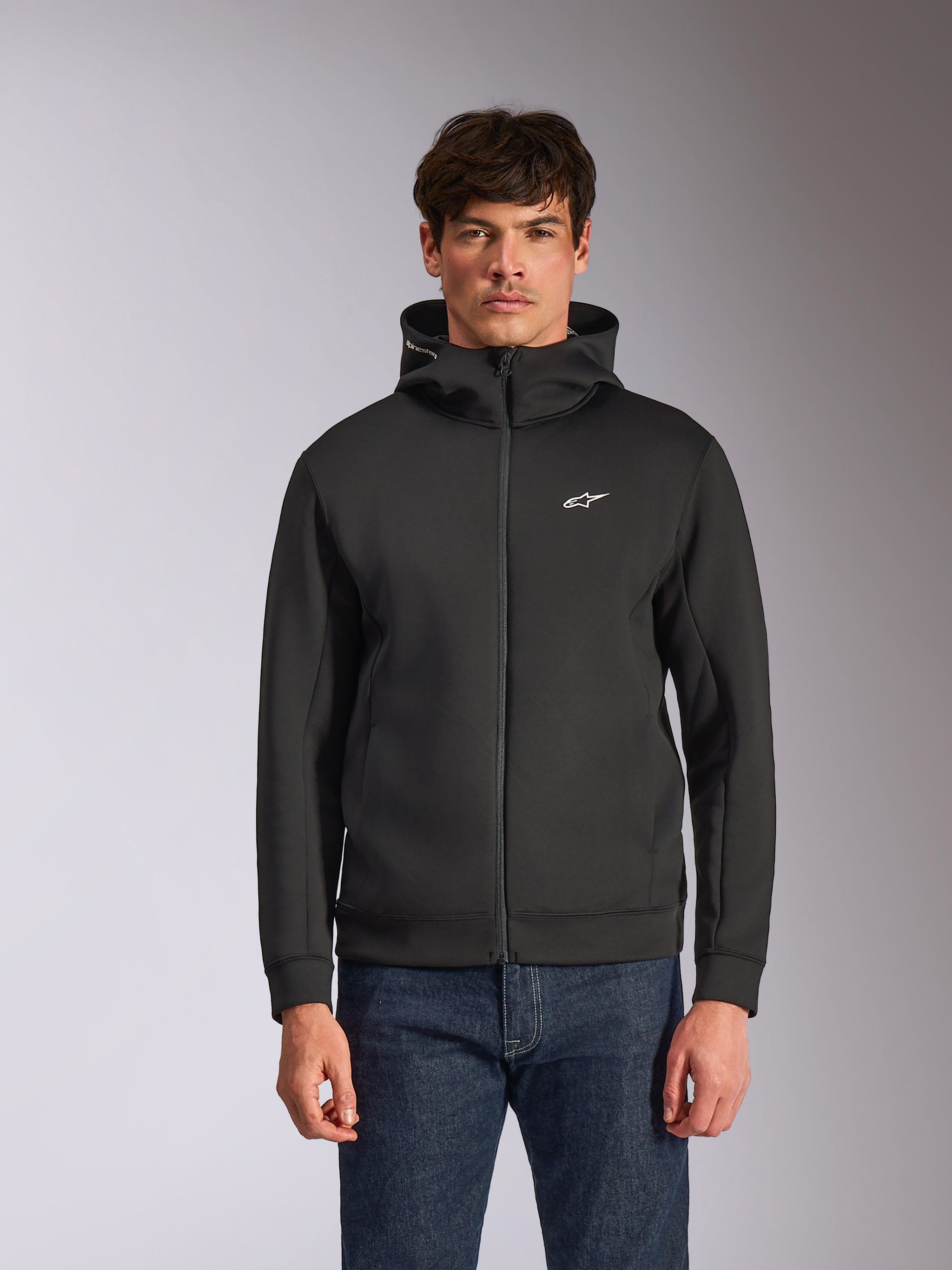 Alpinestars Cypher Hoodie, felpa in pile nera con zip frontale, caratterizzata da un cappuccio a collo alto con branding bianco e un piccolo logo Astars bianco sul petto, indossata da un modello con jeans blu, streetwear casual ispirato al motorsport