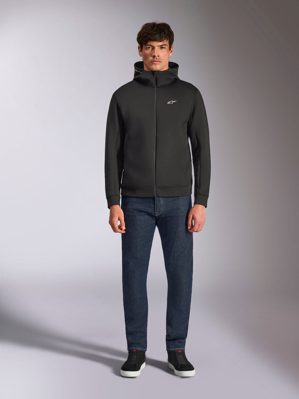 Modello con felpa Alpinestars Cypher Hoodie, felpa in pile nera con zip frontale, cappuccio a collo alto e piccolo logo Astars bianco sul petto, abbinata a jeans scuri e sneakers nere, stile streetwear casual ispirato al motorsport