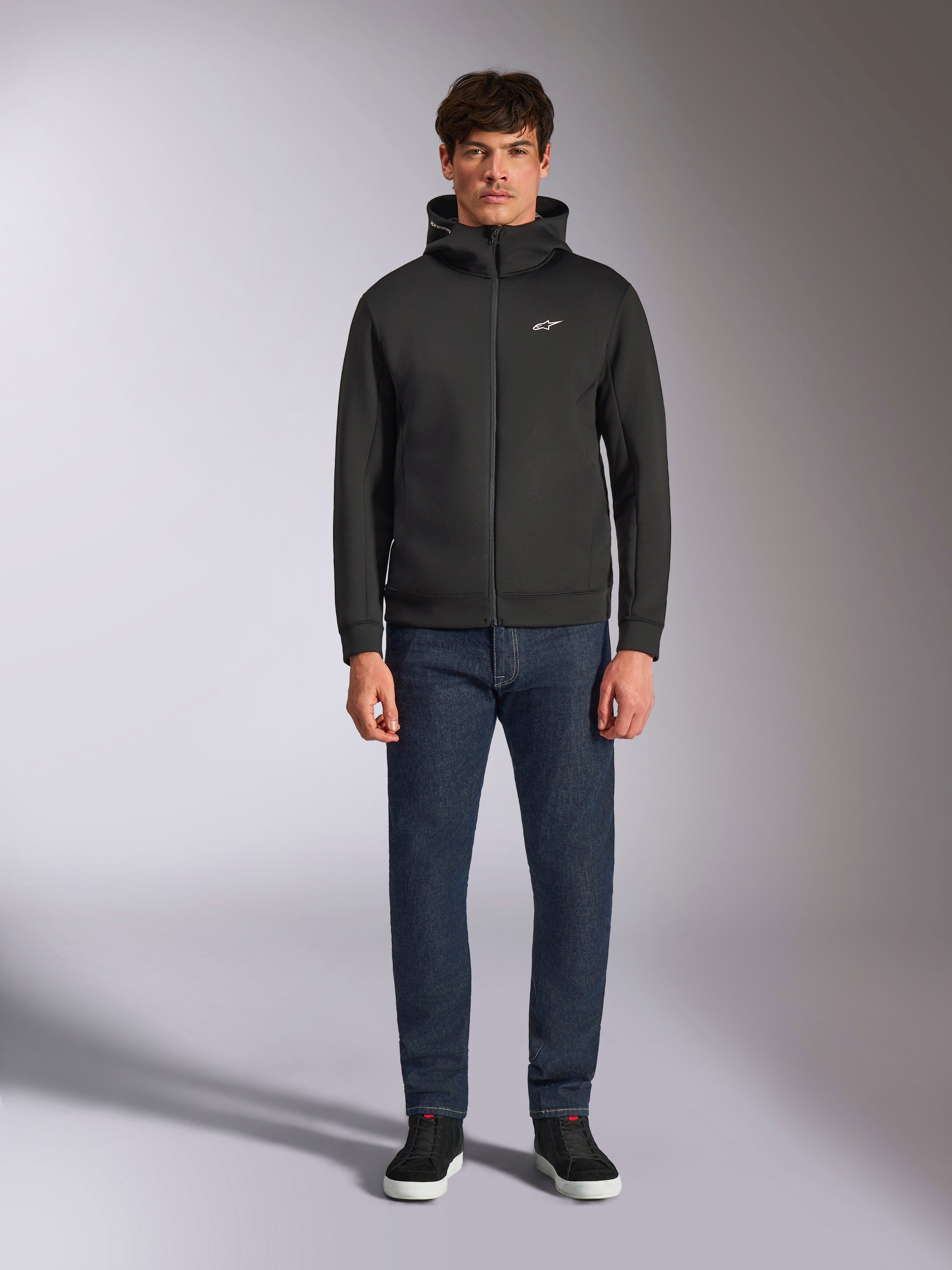 Modello con felpa Alpinestars Cypher Hoodie, felpa in pile nera con zip frontale, cappuccio a collo alto e piccolo logo Astars bianco sul petto, abbinata a jeans scuri e sneakers nere, stile streetwear casual ispirato al motorsport