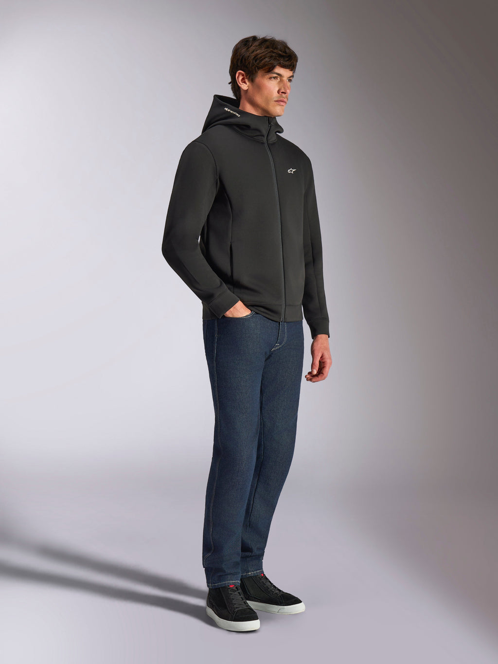 Alpinestars Cypher Hoodie, streetwear casual, felpa nera con zip, modello maschile che la indossa con jeans blu scuro e sneakers nere, cappuccio a collo alto con logo Alpinestars Word, piccolo logo Astars bianco sul petto, look lifestyle elegante ispirato al motorsport