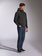 Alpinestars Cypher Hoodie, streetwear casual, felpa nera con zip, modello maschile che la indossa con jeans blu scuro e sneakers nere, cappuccio a collo alto con logo Alpinestars Word, piccolo logo Astars bianco sul petto, look lifestyle elegante ispirato al motorsport