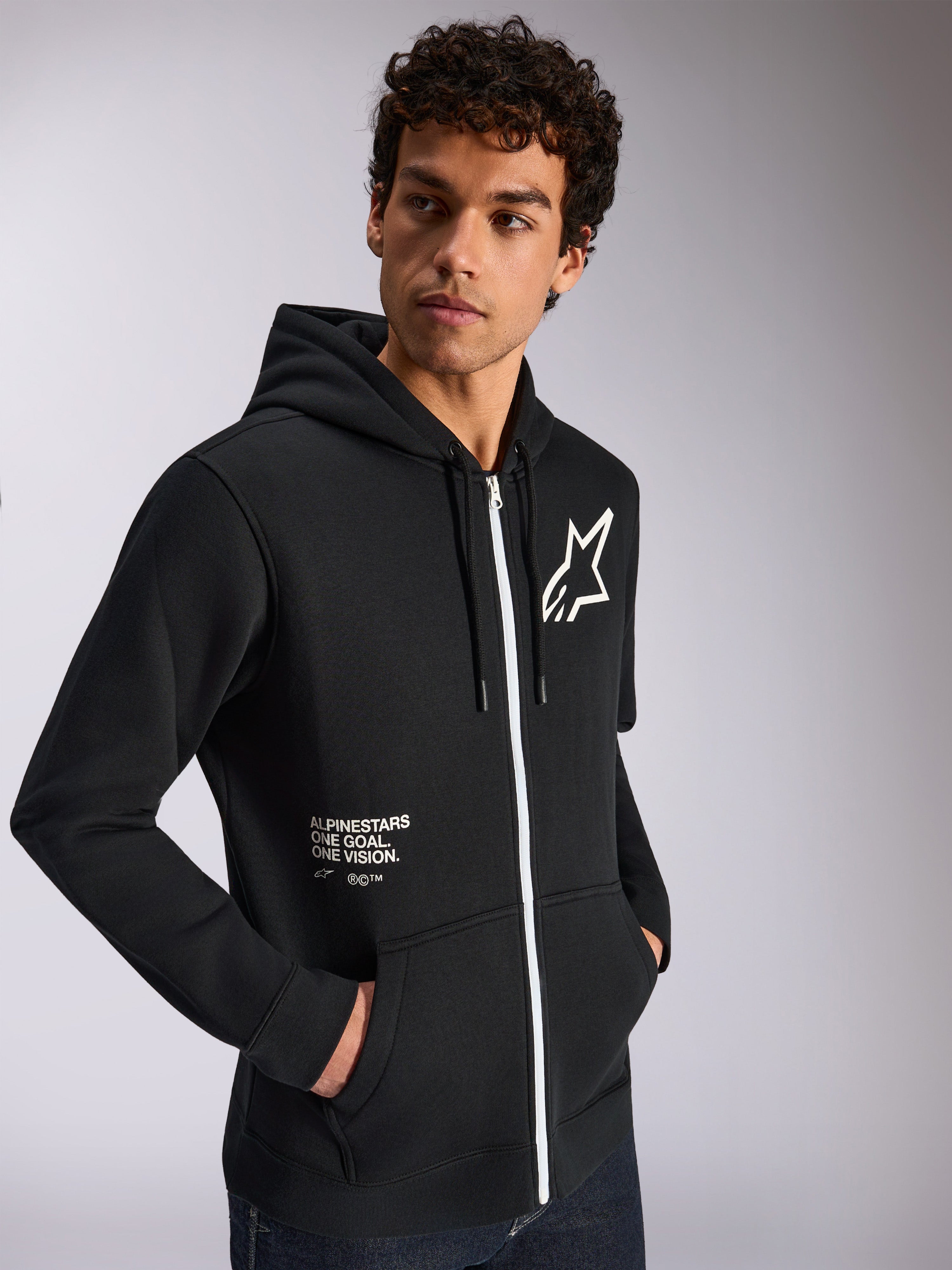 Alpinestars Felpa con cappuccio Transverse, abbigliamento sportivo casual, nero con dettaglio zip bianco, cappuccio con cordino, logo Alpinestars sul petto, grafica 'One Goal One Vision', indossata da un modello con capelli scuri e ricci