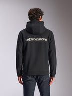 Felpa con cappuccio Alpinestars Transverse, casual streetwear, nera con logo Alpinestars bianco sul retro, felpa in pile con cappuccio e zip frontale, maniche lunghe, indossata da un modello con jeans in denim scuro, design moderno ispirato al motorsport