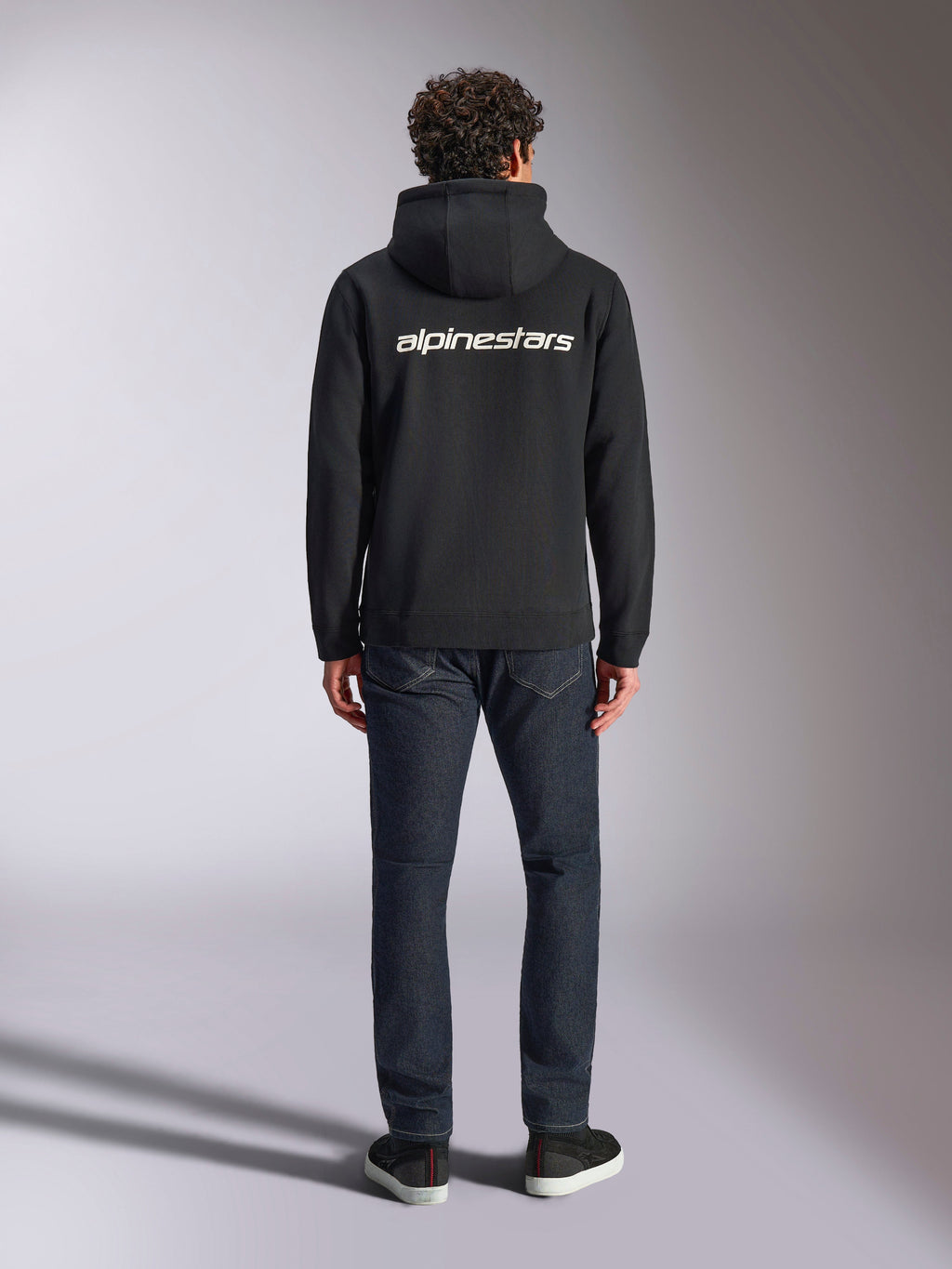 Alpinestars Transverse Hoodie, felpa con cappuccio streetwear casual motorsport, nera con logo Alpinestars bianco sul retro, maniche lunghe, polsini e orlo a coste, vista da dietro con jeans in denim scuro