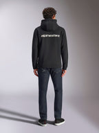 Alpinestars Transverse Hoodie, felpa con cappuccio streetwear casual motorsport, nera con logo Alpinestars bianco sul retro, maniche lunghe, polsini e orlo a coste, vista da dietro con jeans in denim scuro