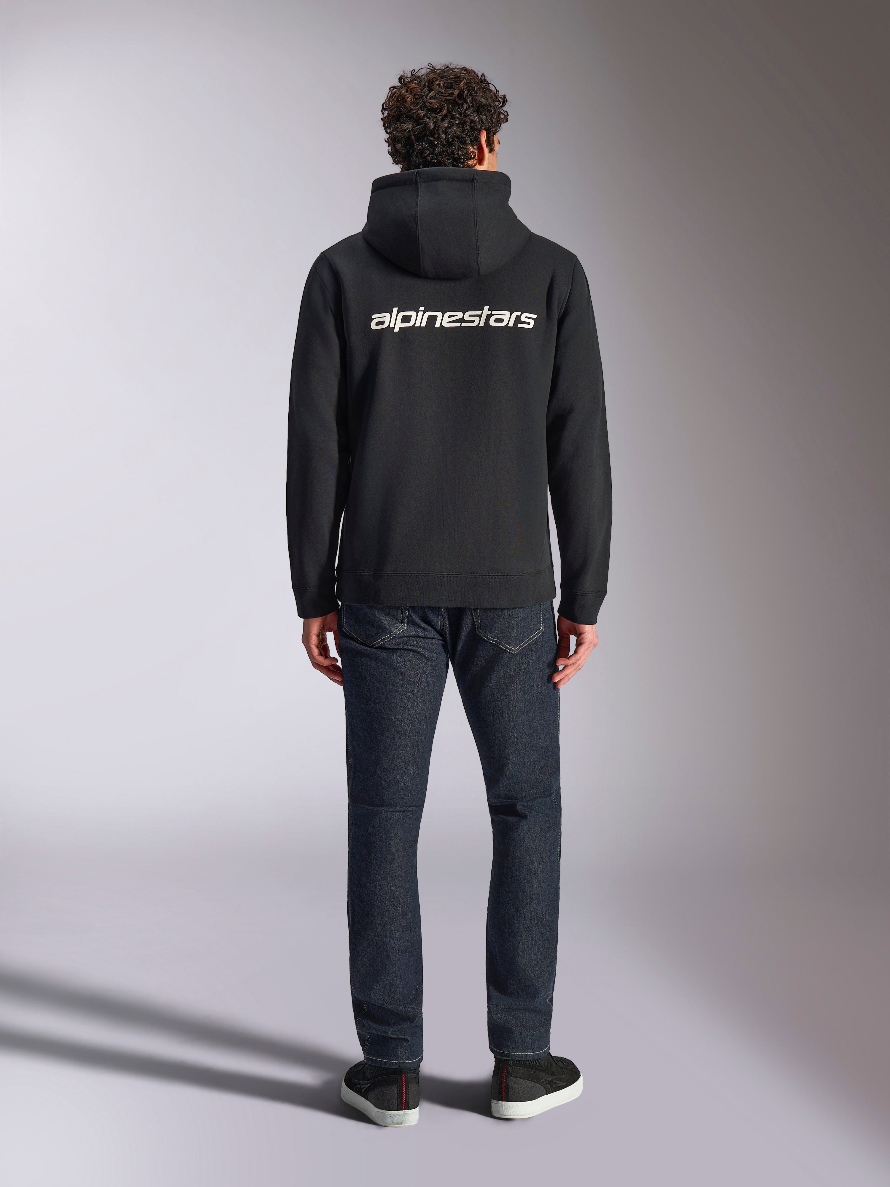 Alpinestars Transverse Hoodie, felpa con cappuccio streetwear casual motorsport, nera con logo Alpinestars bianco sul retro, maniche lunghe, polsini e orlo a coste, vista da dietro con jeans in denim scuro