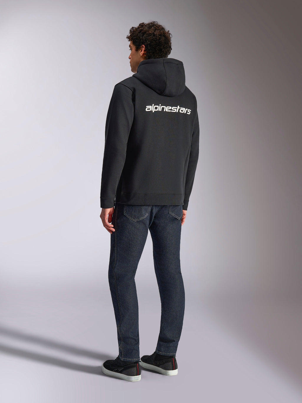 Felpa con cappuccio Alpinestars Transverse, felpa nera con zip frontale, vista posteriore con logo Alpinestars bianco sulle spalle, indossata con jeans blu scuro e sneakers nere, streetwear casual ispirato al motorsport