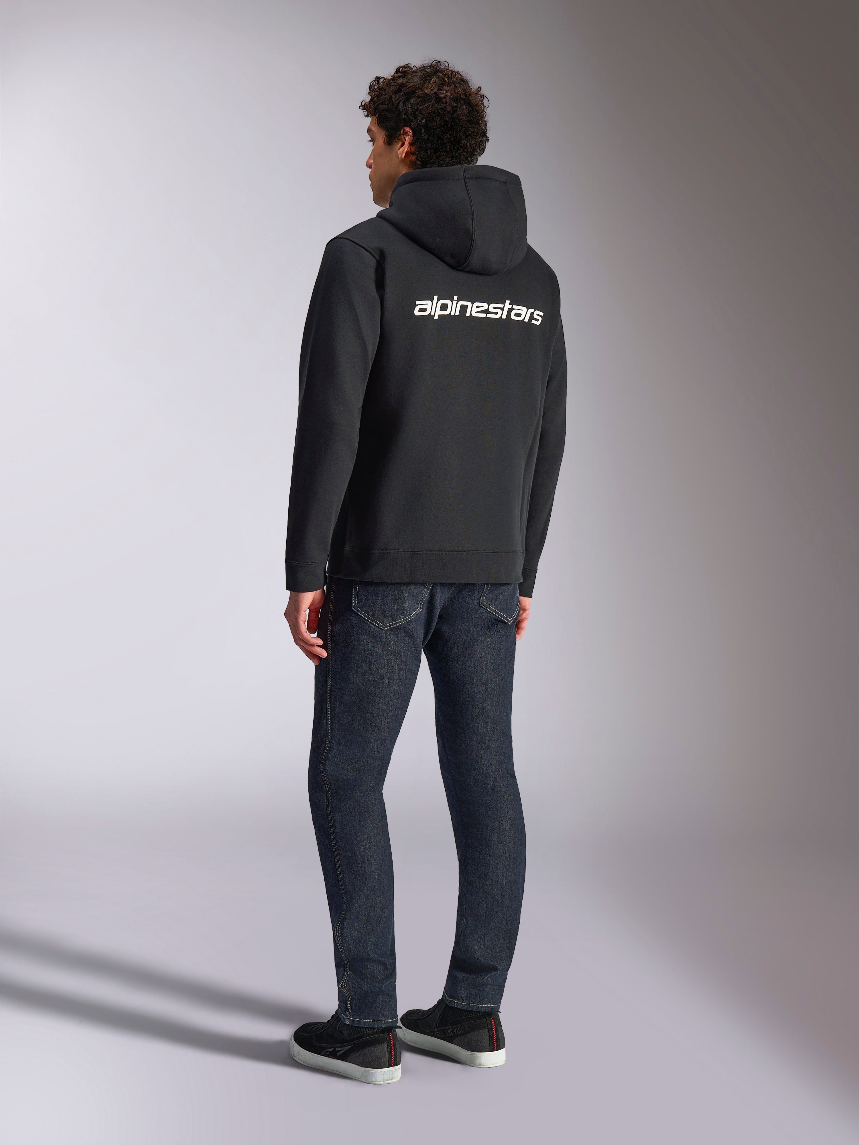 Felpa con cappuccio Alpinestars Transverse, felpa nera con zip frontale, vista posteriore con logo Alpinestars bianco sulle spalle, indossata con jeans blu scuro e sneakers nere, streetwear casual ispirato al motorsport