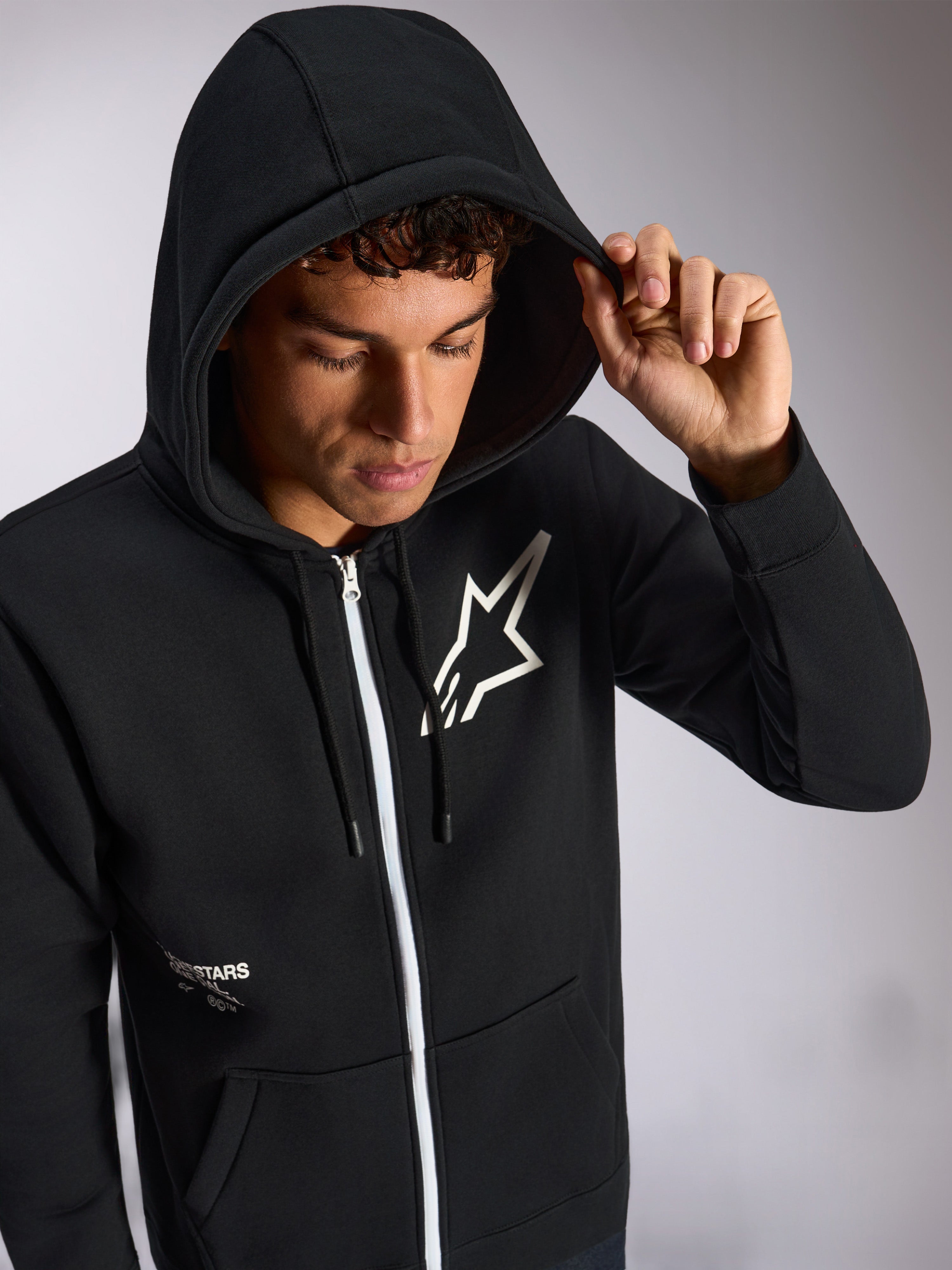 Alpinestars Transverse Hoodie, felpa nera con zip frontale, logo Astars bianco sul petto, zip bianca, cappuccio con cordino indossato, streetwear casual ispirato al motorsport