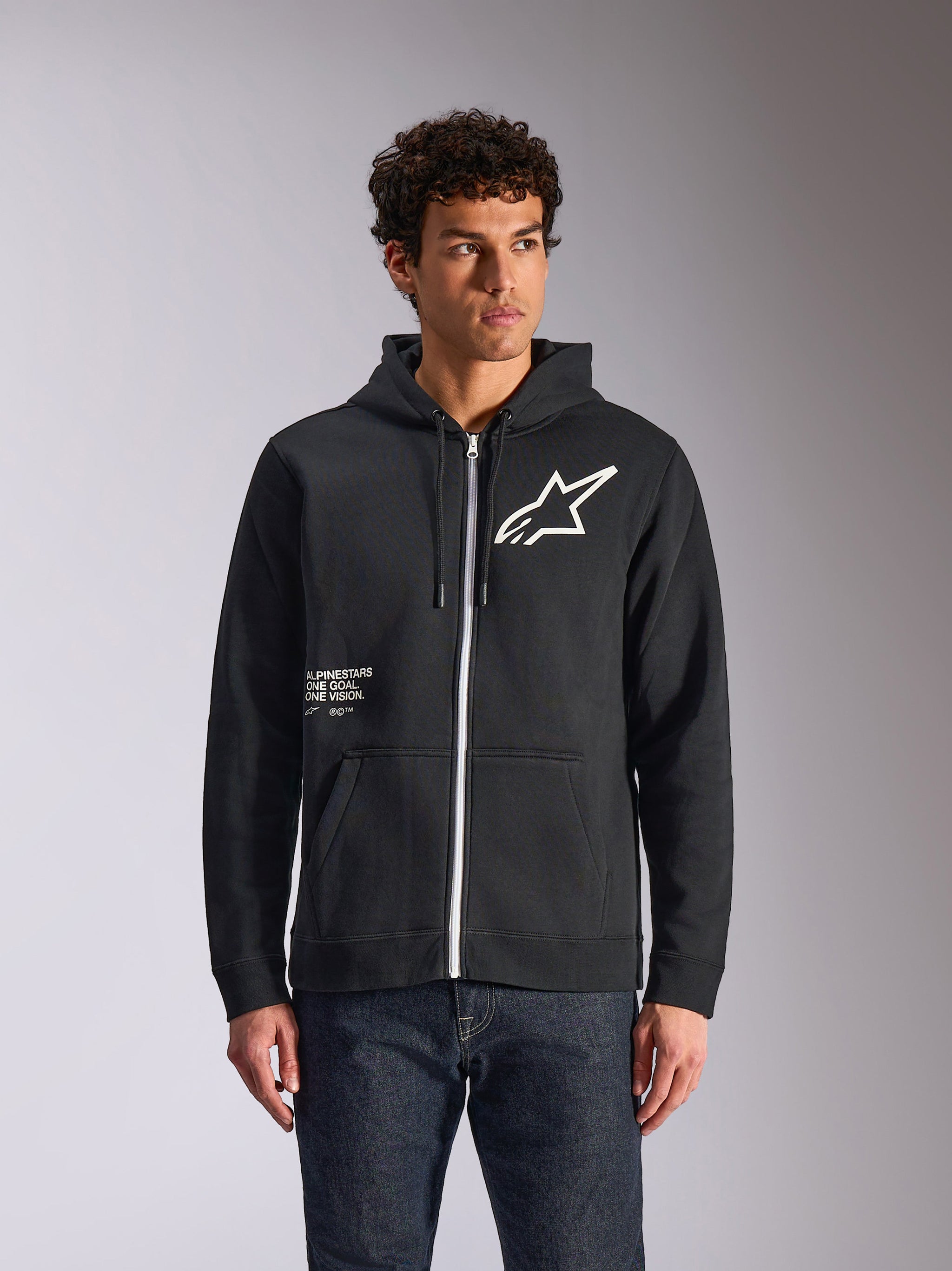 Alpinestars Transverse Hoodie, felpa nera con cappuccio e zip frontale, logo Astars bianco e grafica One Goal sul davanti, indossata da un modello con jeans scuri, abbigliamento streetwear casual ispirato al motorsport