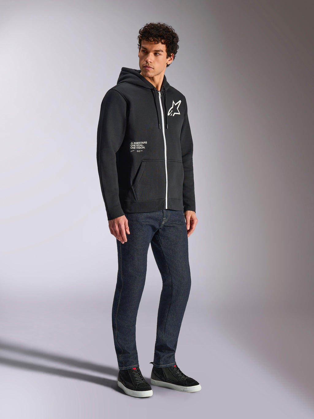 Alpinestars Transverse Hoodie, felpa nera con cappuccio e zip bianca a contrasto, logo a stella bianco sul petto e scritta 'One Goal' sulla tasca, indossata da un modello con jeans scuri e sneakers nere, streetwear casual ispirato al motorsport