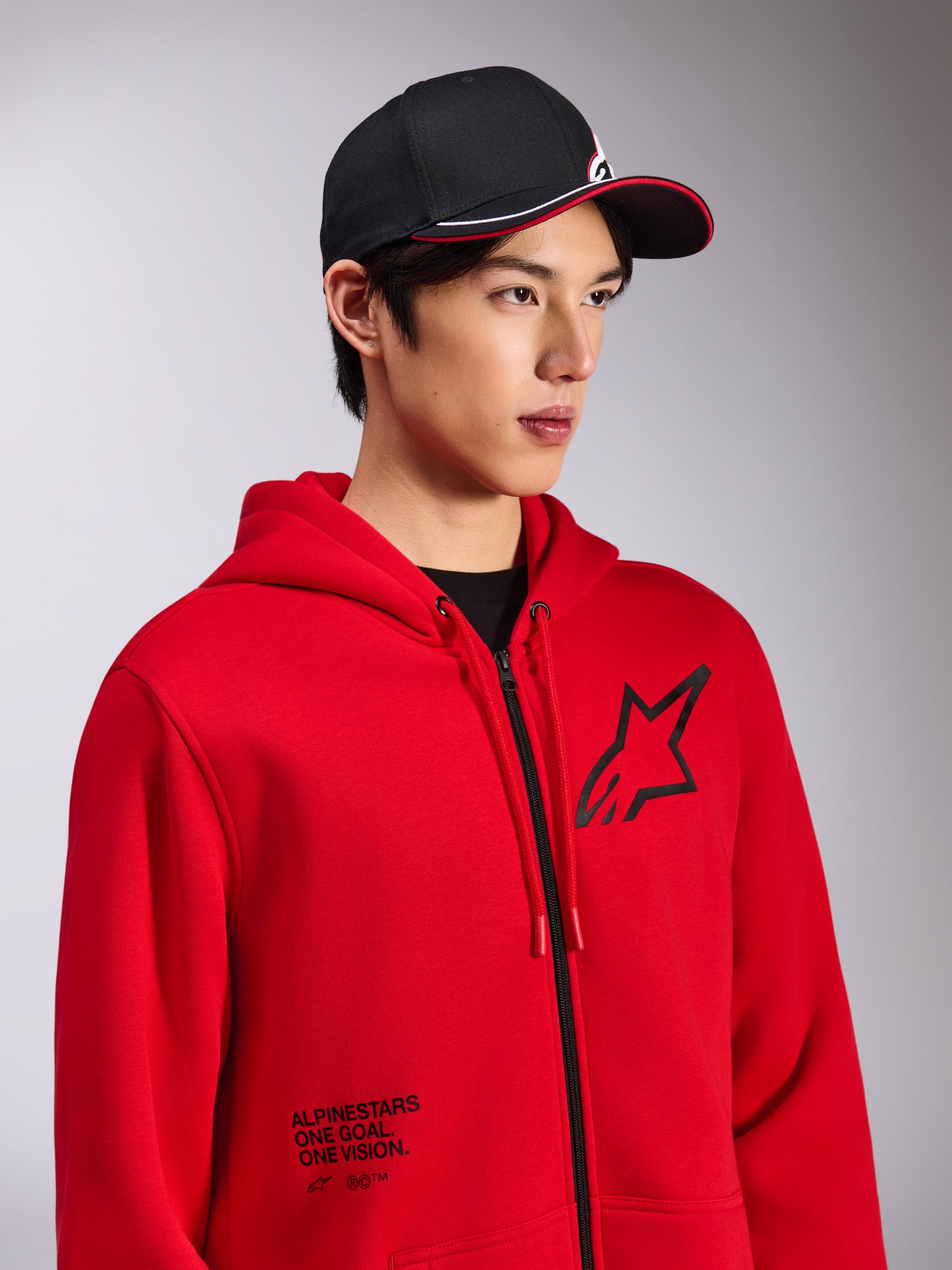 Alpinestars Transverse Hoodie, felpa rossa con zip frontale, logo Astars nero sul petto e testo 'One Goal. One Vision.' sulla parte inferiore frontale, streetwear casual motorsport, indossata da un modello con cappellino nero
