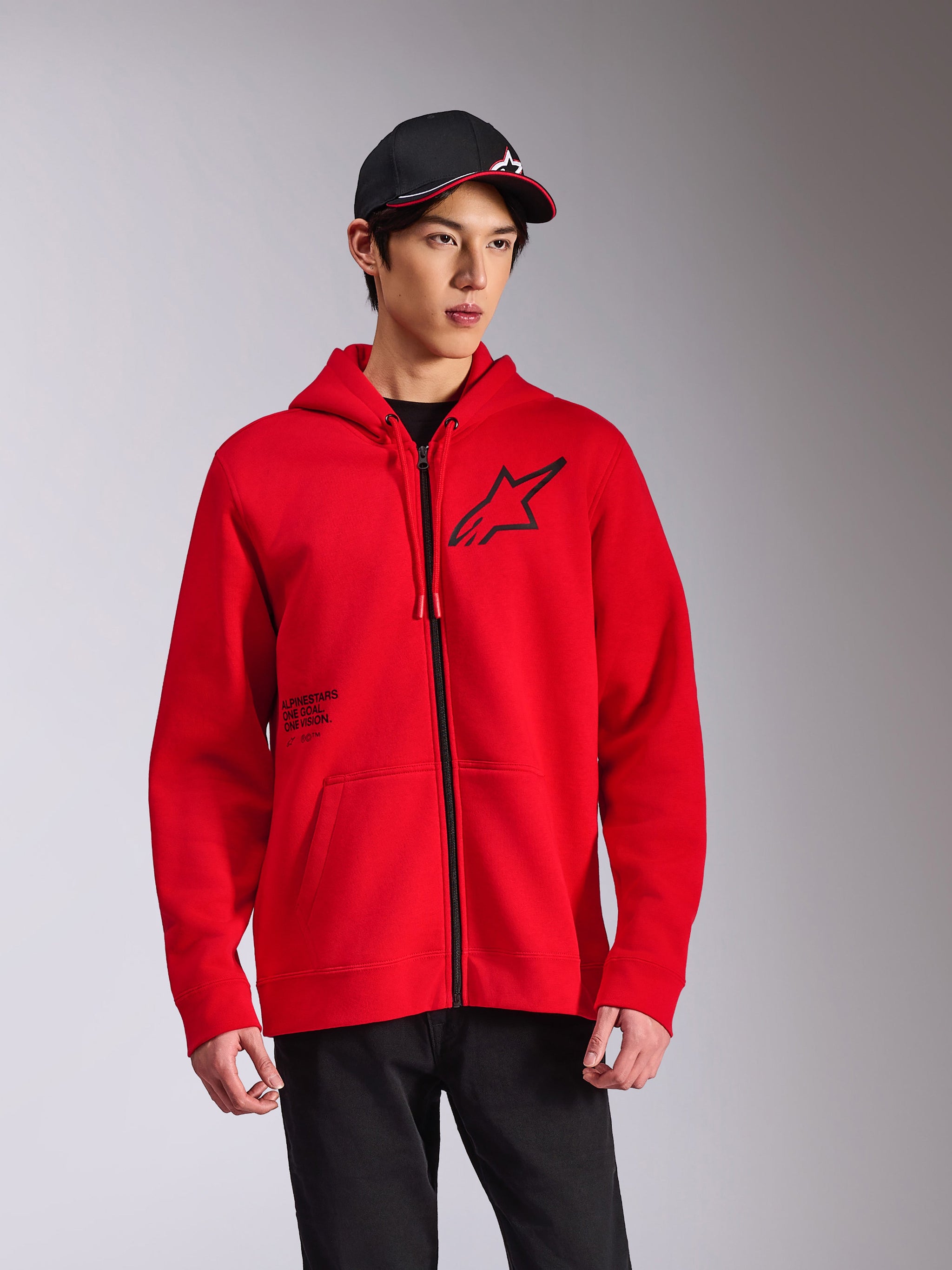 Alpinestars Transverse Hoodie, felpa in pile rossa con zip frontale, logo Astars nero sul petto e stampa 'One Goal', stile streetwear casual indossato con cappellino e pantaloni neri