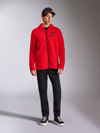 Alpinestars Transverse Hoodie, felpa in pile rossa con zip integrale, logo Astars nero sul petto e stampa One Goal sul lato, indossata da un modello con pantaloni e cappellino neri, streetwear casual ispirato al motorsport