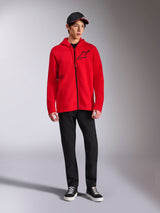 Alpinestars Transverse Hoodie, felpa in pile rossa con zip integrale, logo Astars nero sul petto e stampa One Goal sul lato, indossata da un modello con pantaloni e cappellino neri, streetwear casual ispirato al motorsport