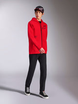 Alpinestars Felpa con cappuccio Transverse in rosso, modello che indossa una felpa con cappuccio e zip integrale, logo Astars nero e scritte stampate, abbinata a pantaloni neri e cappellino, streetwear casual motorsport