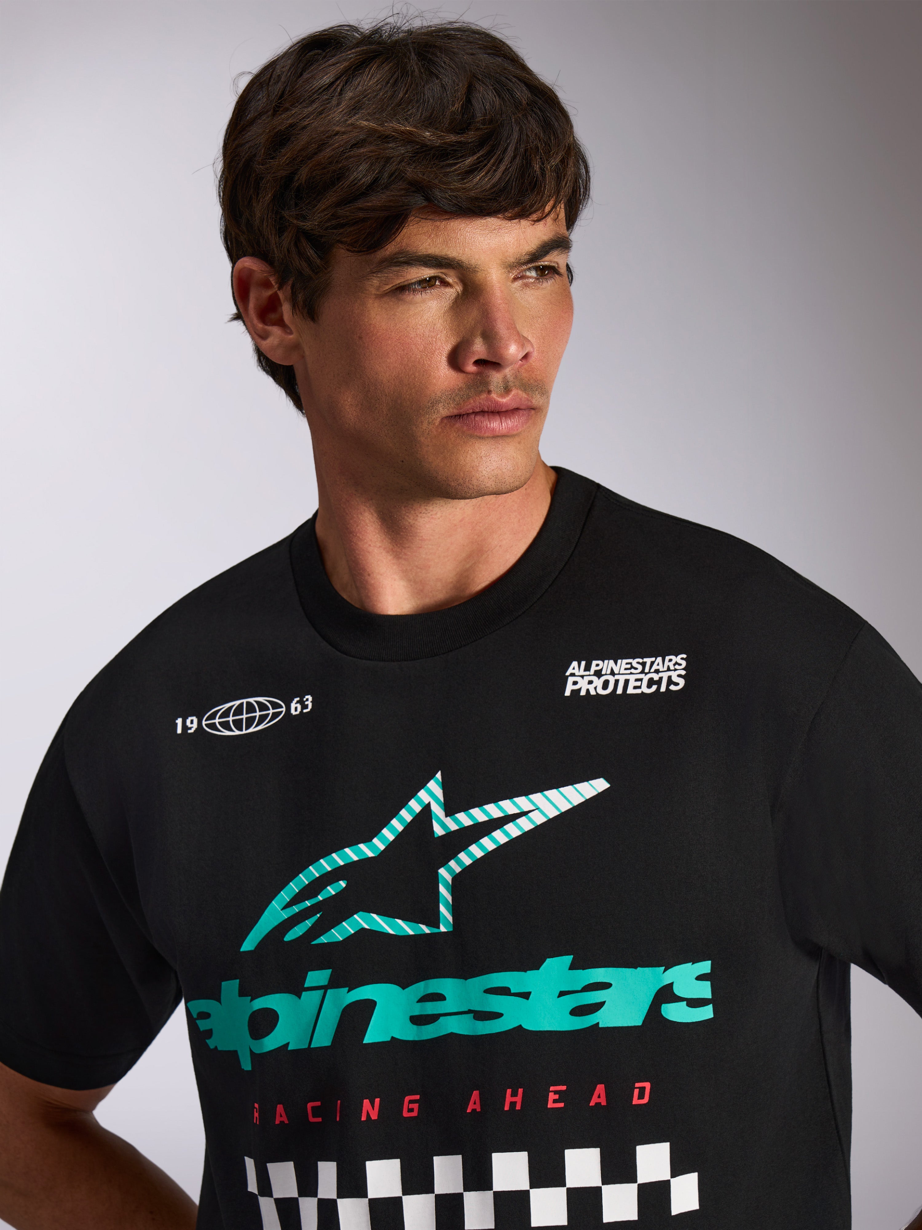 Alpinestars Aplomb Oversized CSF Tee, t-shirt a maniche corte, nero, logo combinato Astars bianco e verde acqua con scritta 'Racing Ahead' e motivo a scacchi, indossata da un modello, streetwear casual motorsport