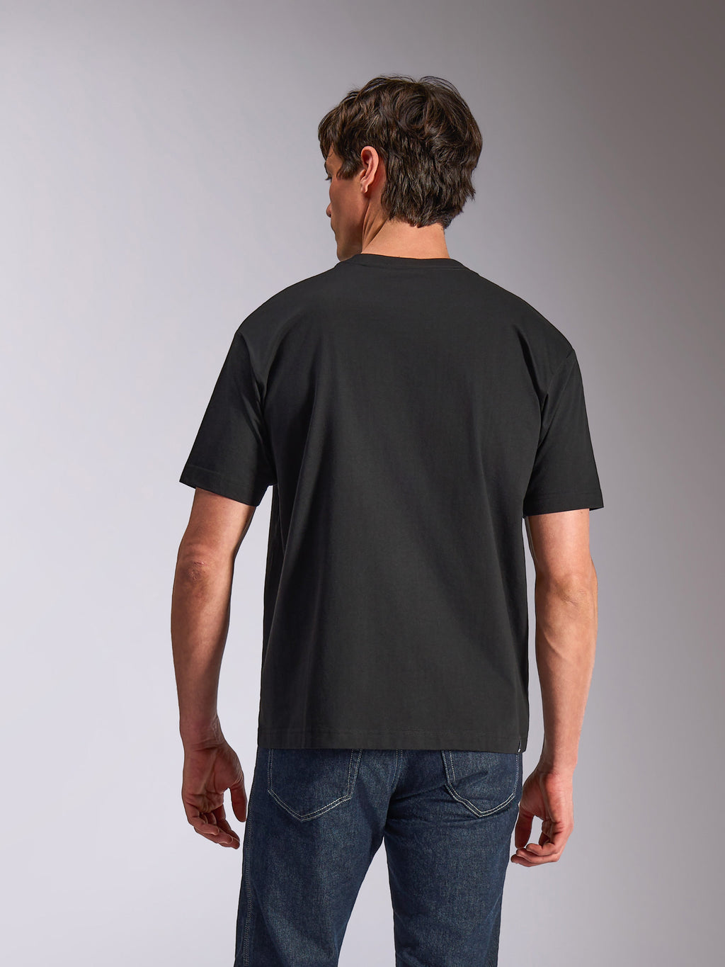 Alpinestars Aplomb Oversized CSF Tee, t-shirt a maniche corte, nero, vista posteriore, indossata da un modello con jeans blu, abbigliamento sportivo casual dalla vestibilità rilassata
