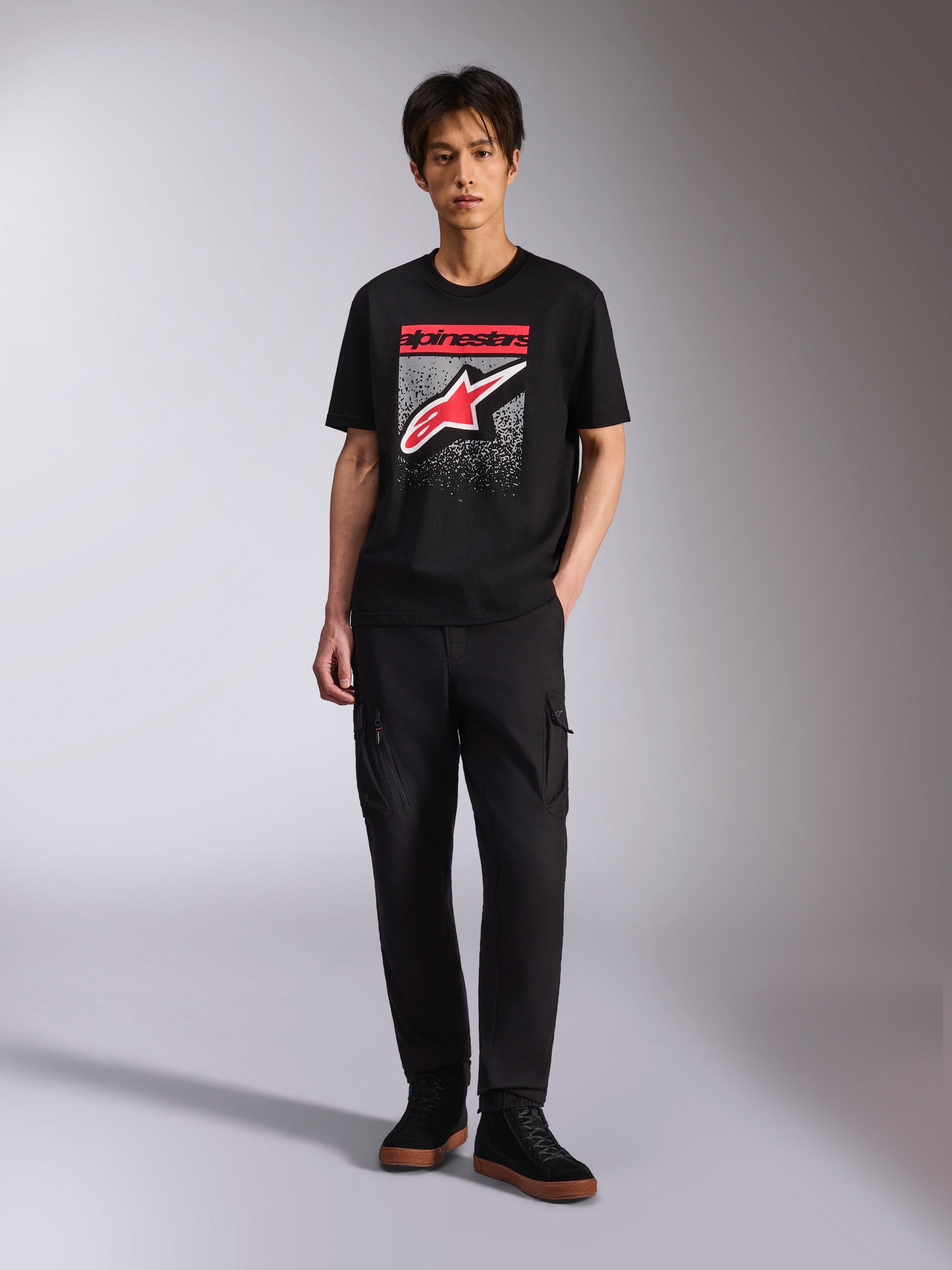 Alpinestars Noka CSF Tee, t-shirt a maniche corte, nero, logo Astars frontale con grafica a gradazione MX rossa e bianca, indossata da un modello con pantaloni cargo neri, streetwear motorsport casual