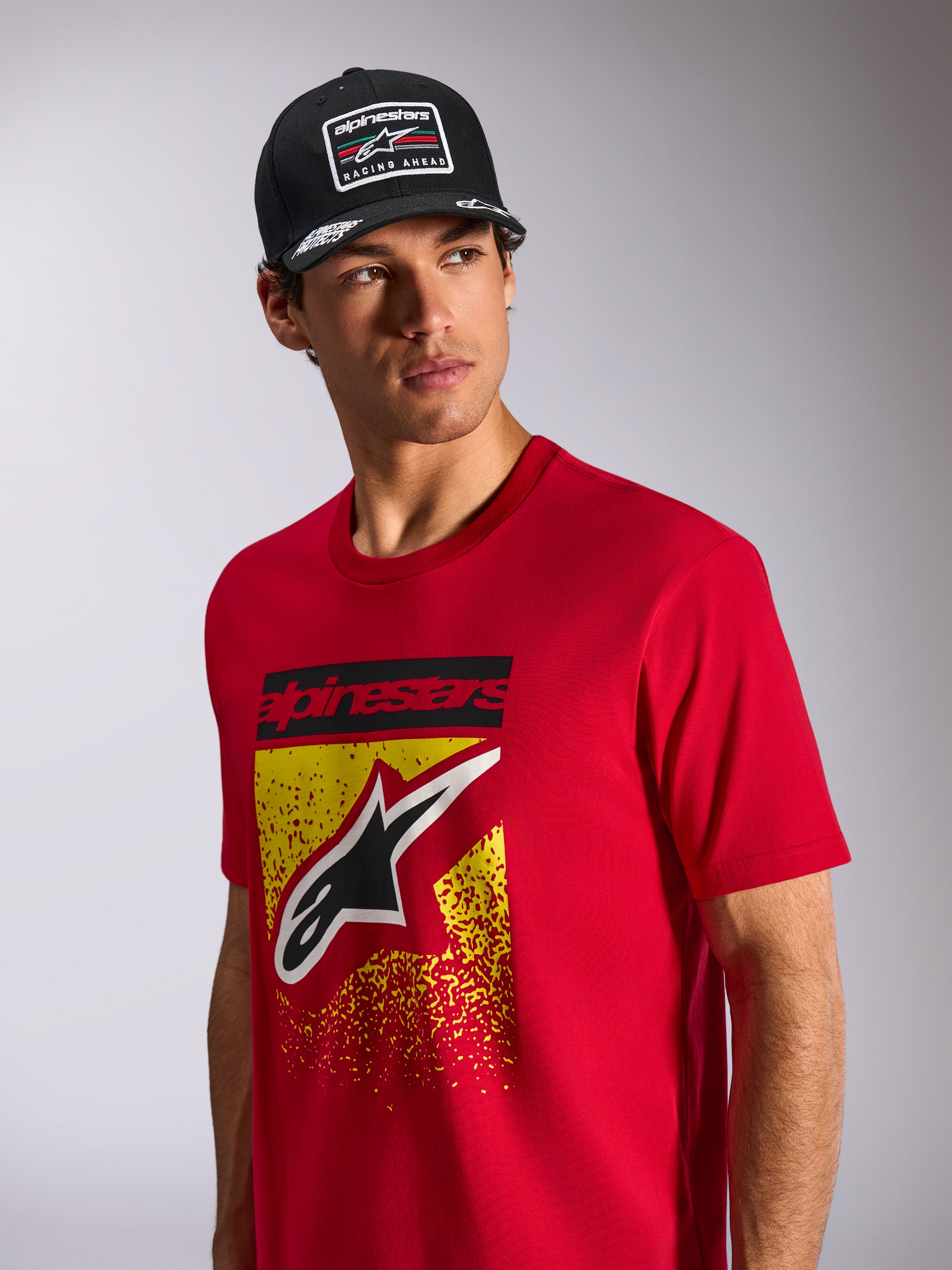 Alpinestars Noka CSF Tee, t-shirt a maniche corte, rossa, con logo Astars frontale con sfumatura MX gialla e rossa, indossata con cappellino Alpinestars 'Racing Ahead' nero, abbigliamento casual motorsport streetwear