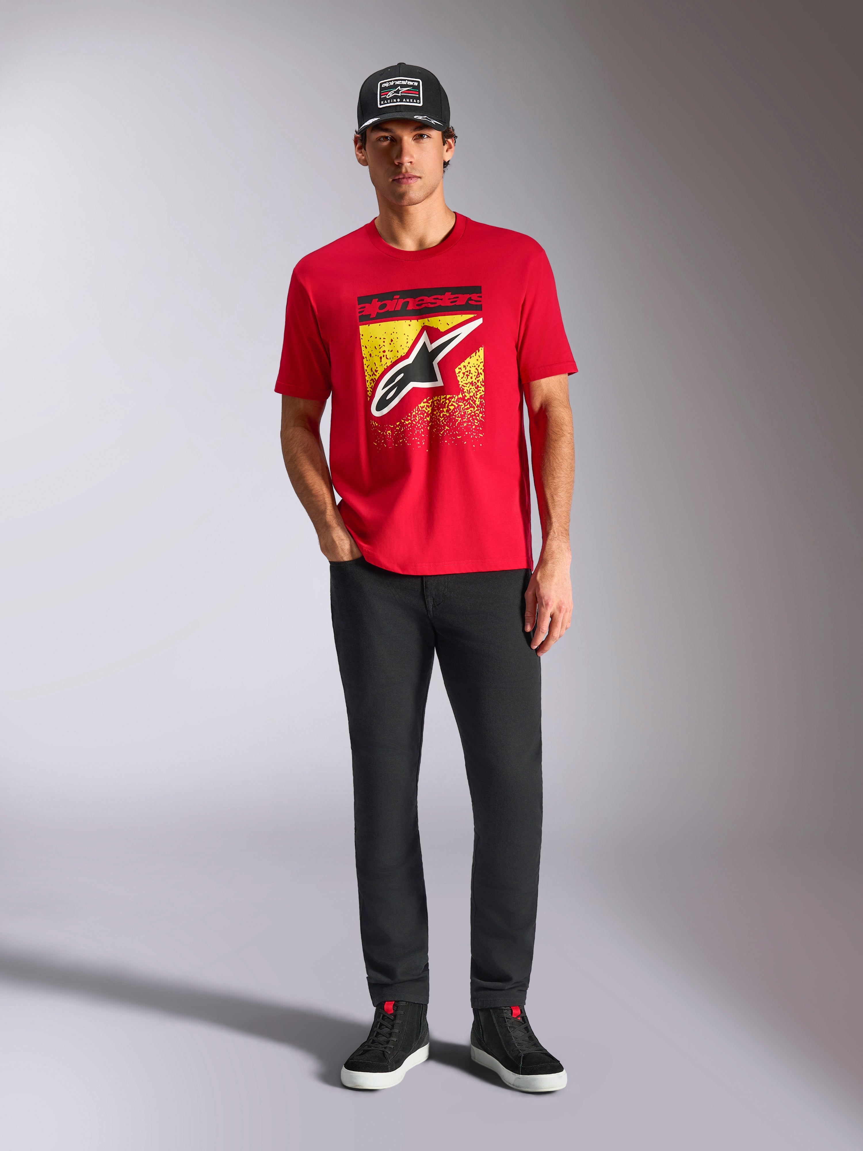 Alpinestars Noka CSF Tee, t-shirt a maniche corte, rossa, con un grande logo Astars frontale con gradazione MX gialla e nera, indossata con pantaloni neri e un cappellino nero, streetwear casual motorsport