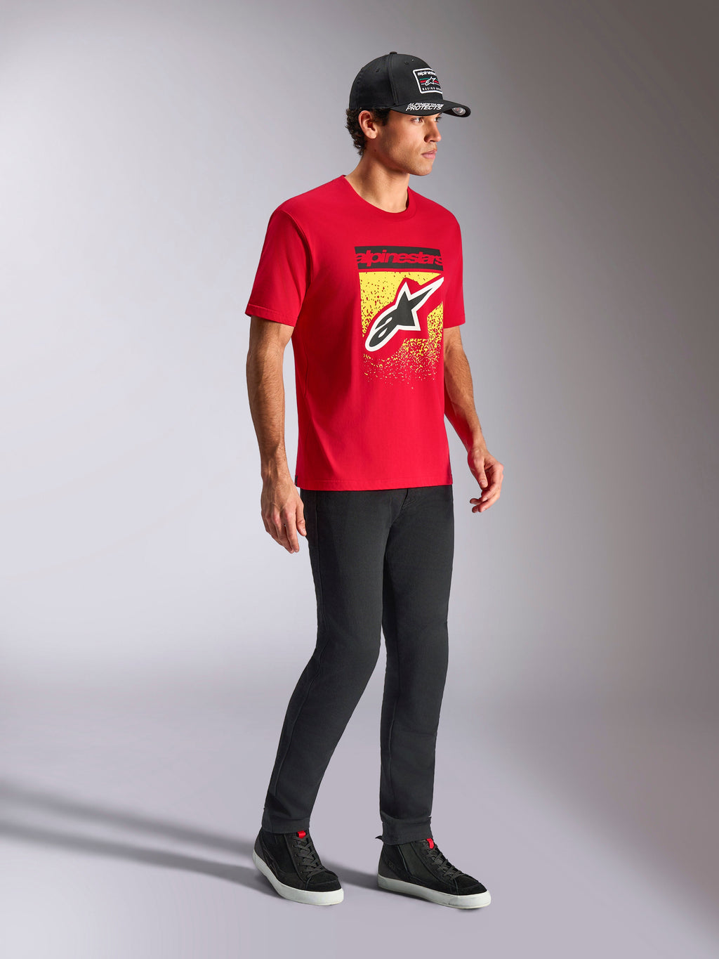 Alpinestars Noka CSF Tee, t-shirt a maniche corte, rosso, con logo Astars frontale e gradazione MX su grafica gialla, indossata con cappellino Alpinestars nero e pantaloni neri, streetwear casual da motorsport