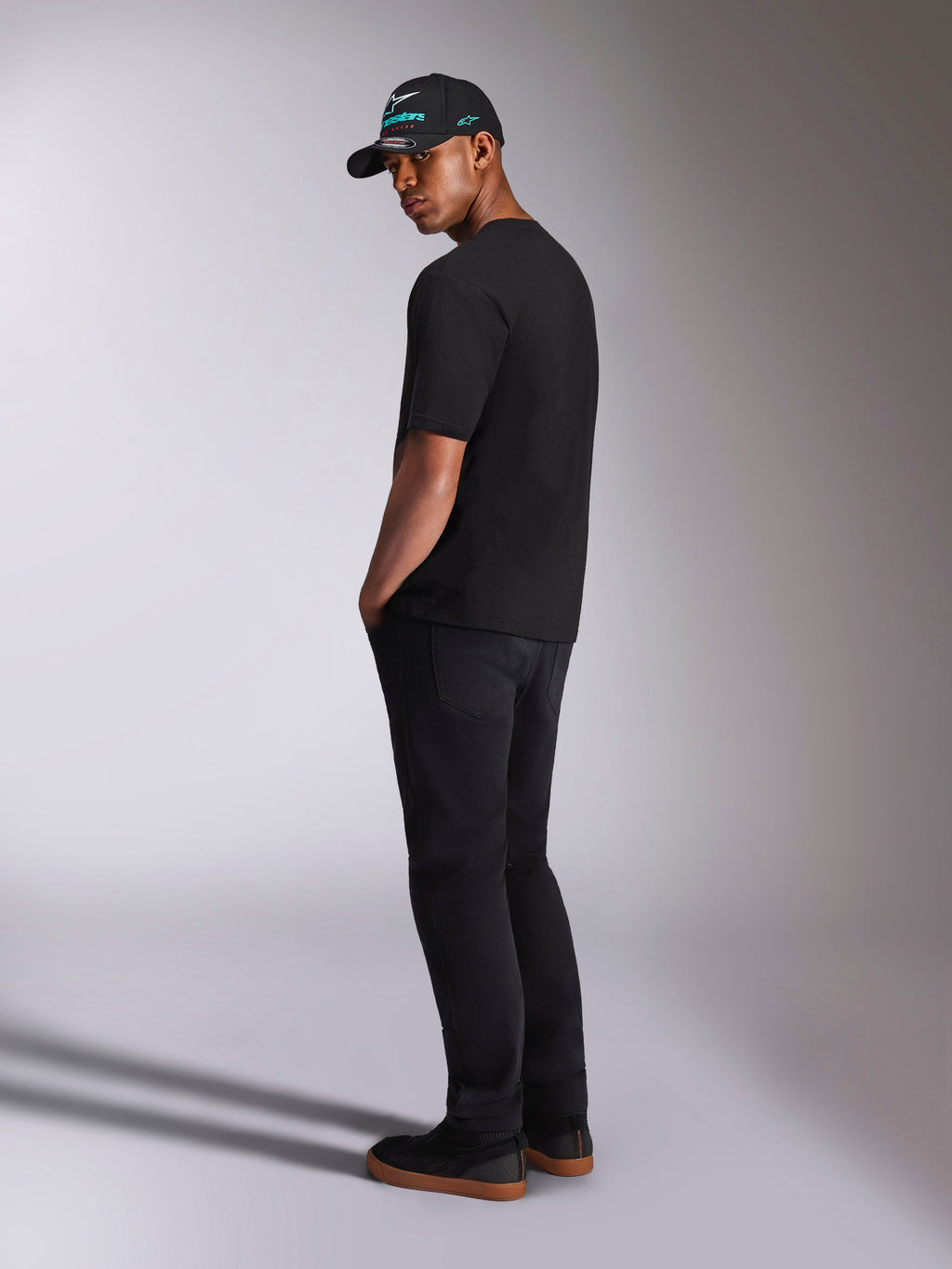 Alpinestars Ellipsoid CSF Tee, maglietta a maniche corte, nera, vista posteriore su modello con pantaloni neri e cappellino Alpinestars nero, outfit casual in stile motorsport