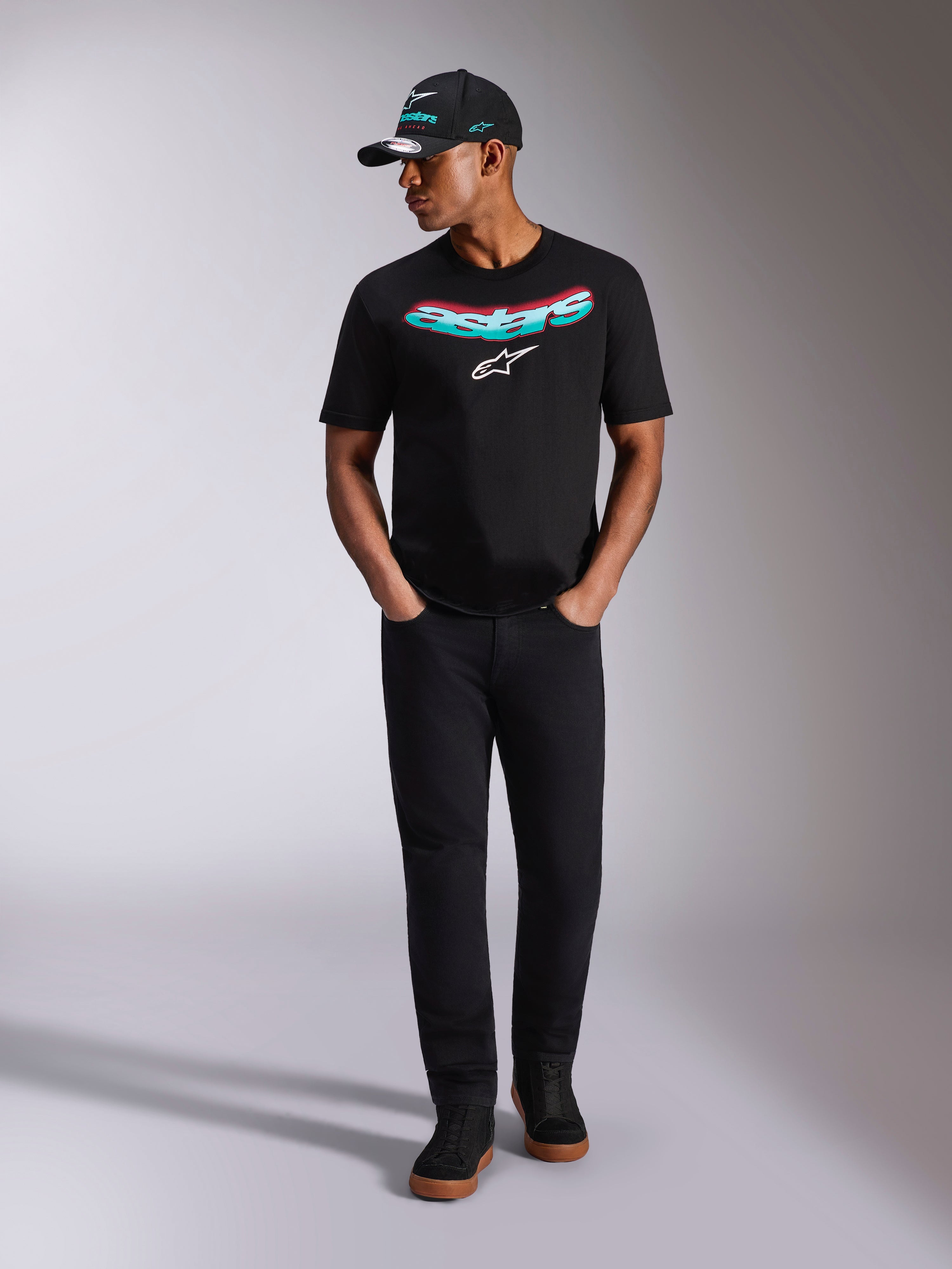 Alpinestars Ellipsoid CSF Tee, maglietta a maniche corte, nera, scritta Astars curva con gradiente verde acqua e rosa e logo bianco, indossata da un modello con cappellino e pantaloni neri coordinati, abbigliamento casual motorsport lifestyle