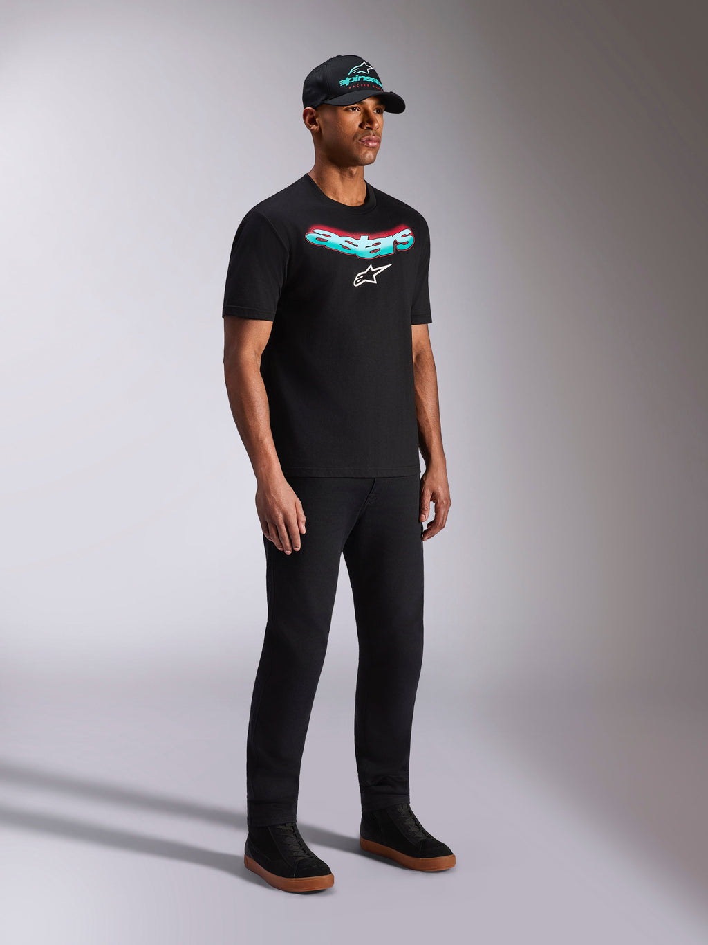 Alpinestars Ellipsoid CSF Tee, maglietta a maniche corte nera, grafica Astars con gradiente verde acqua e rosso e logo bianco, modello che indossa abbigliamento casual streetwear con cappellino e pantaloni neri
