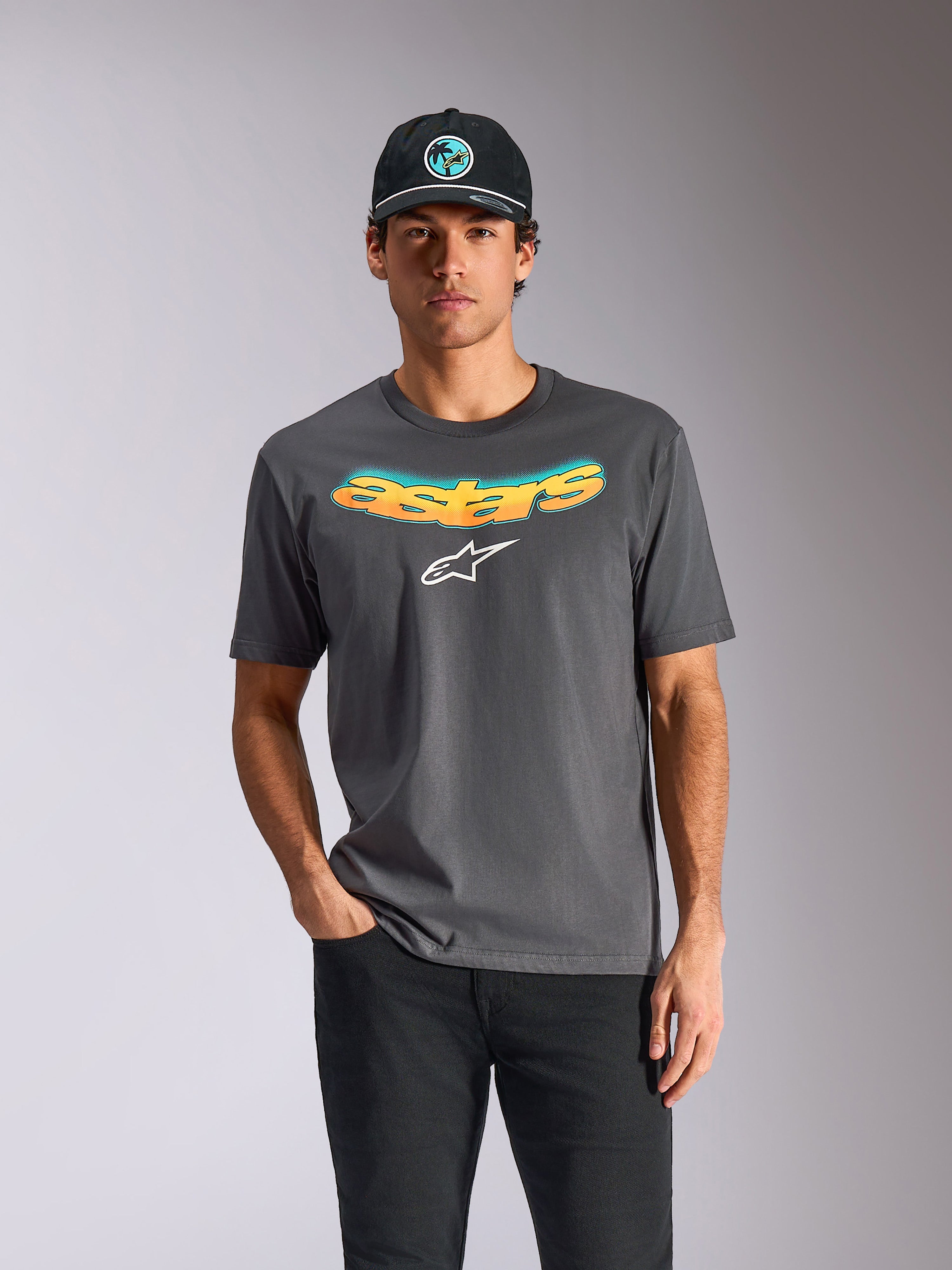 Alpinestars Ellipsoid CSF Tee, t-shirt a maniche corte, grigio antracite, scritta 'astars' curva con gradiente arancione e blu e un logo bianco, indossata da un modello con berretto e pantaloni neri, streetwear casual motorsport