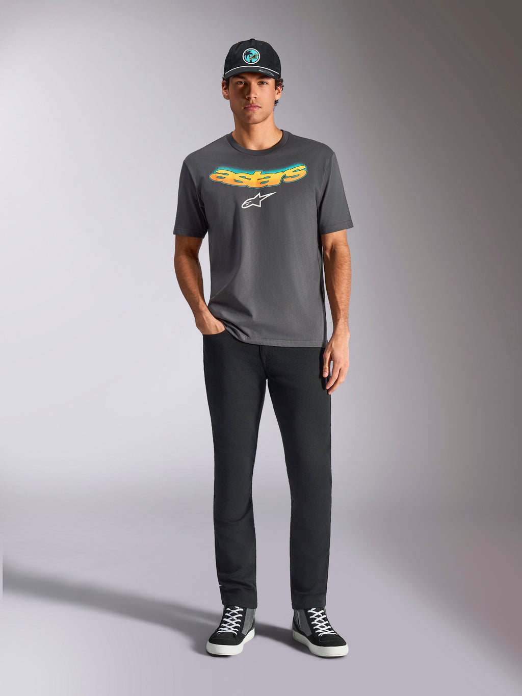 Alpinestars Ellipsoid CSF Tee, t-shirt a maniche corte, grigio antracite, con scritta curva a gradiente giallo e arancione e logo Astars bianco, indossata da un modello con pantaloni neri e cappellino da baseball, stile casual sportswear