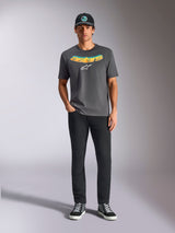Alpinestars Ellipsoid CSF Tee, t-shirt a maniche corte, grigio antracite, con scritta curva a gradiente giallo e arancione e logo Astars bianco, indossata da un modello con pantaloni neri e cappellino da baseball, stile casual sportswear