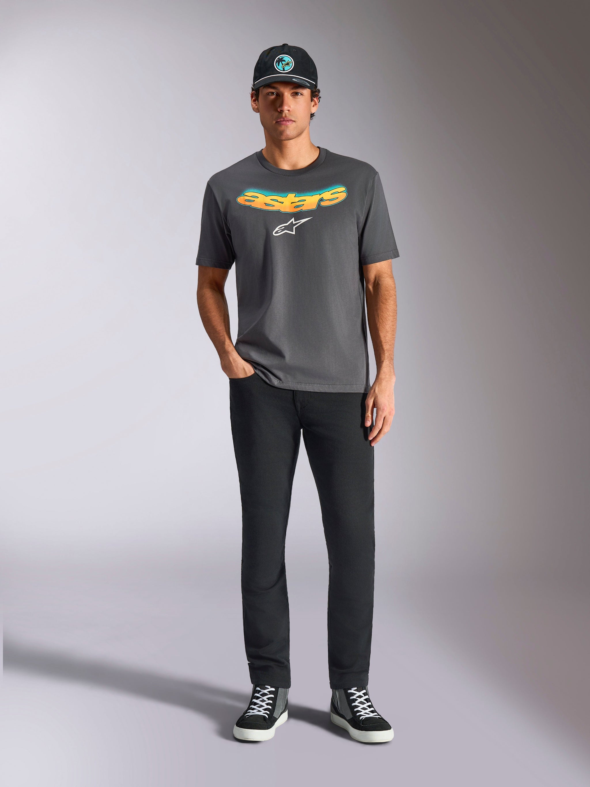 Alpinestars Ellipsoid CSF Tee, t-shirt a maniche corte, grigio antracite, con scritta curva a gradiente giallo e arancione e logo Astars bianco, indossata da un modello con pantaloni neri e cappellino da baseball, stile casual sportswear