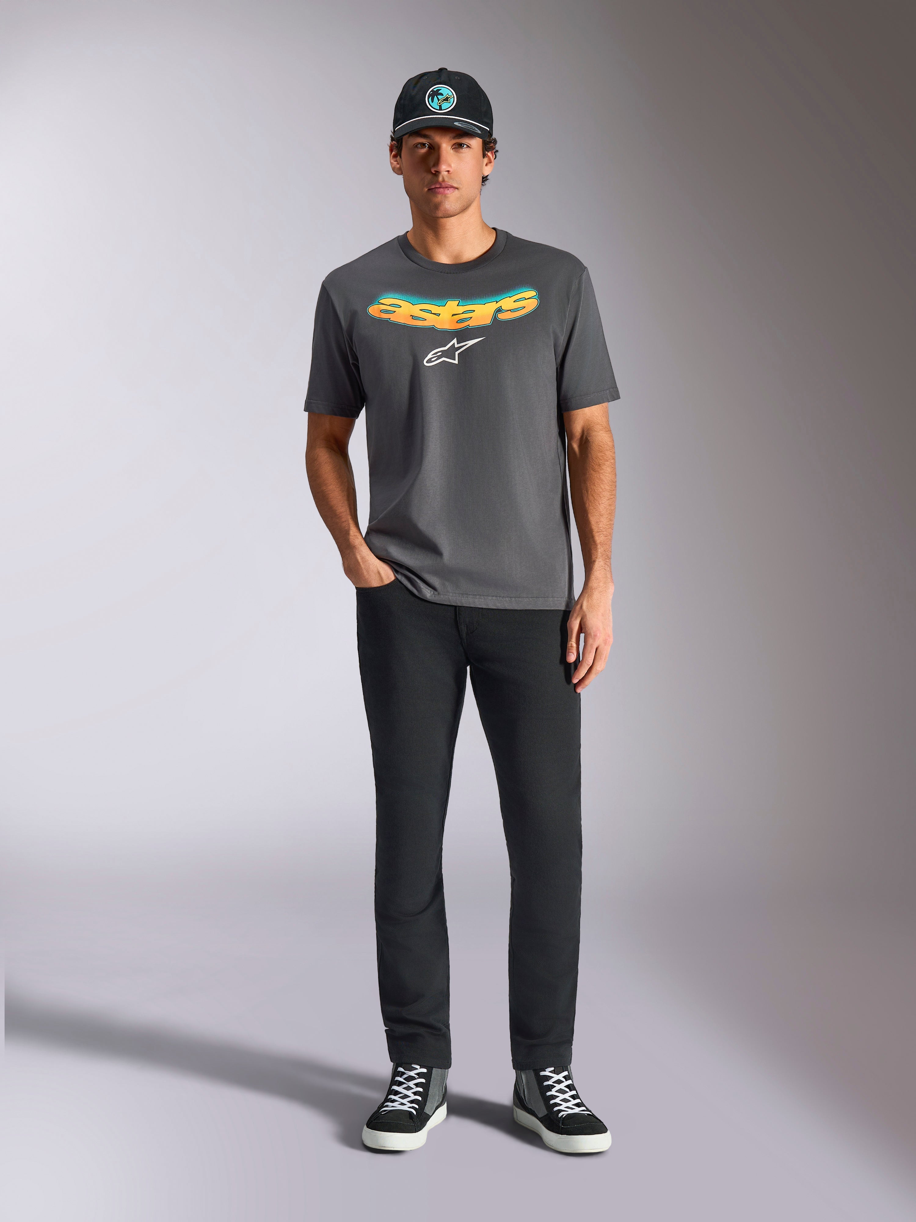Alpinestars Ellipsoid CSF Tee, t-shirt a maniche corte, grigio antracite, con scritta curva a gradiente giallo e arancione e logo Astars bianco, indossata da un modello con pantaloni neri e cappellino da baseball, stile casual sportswear