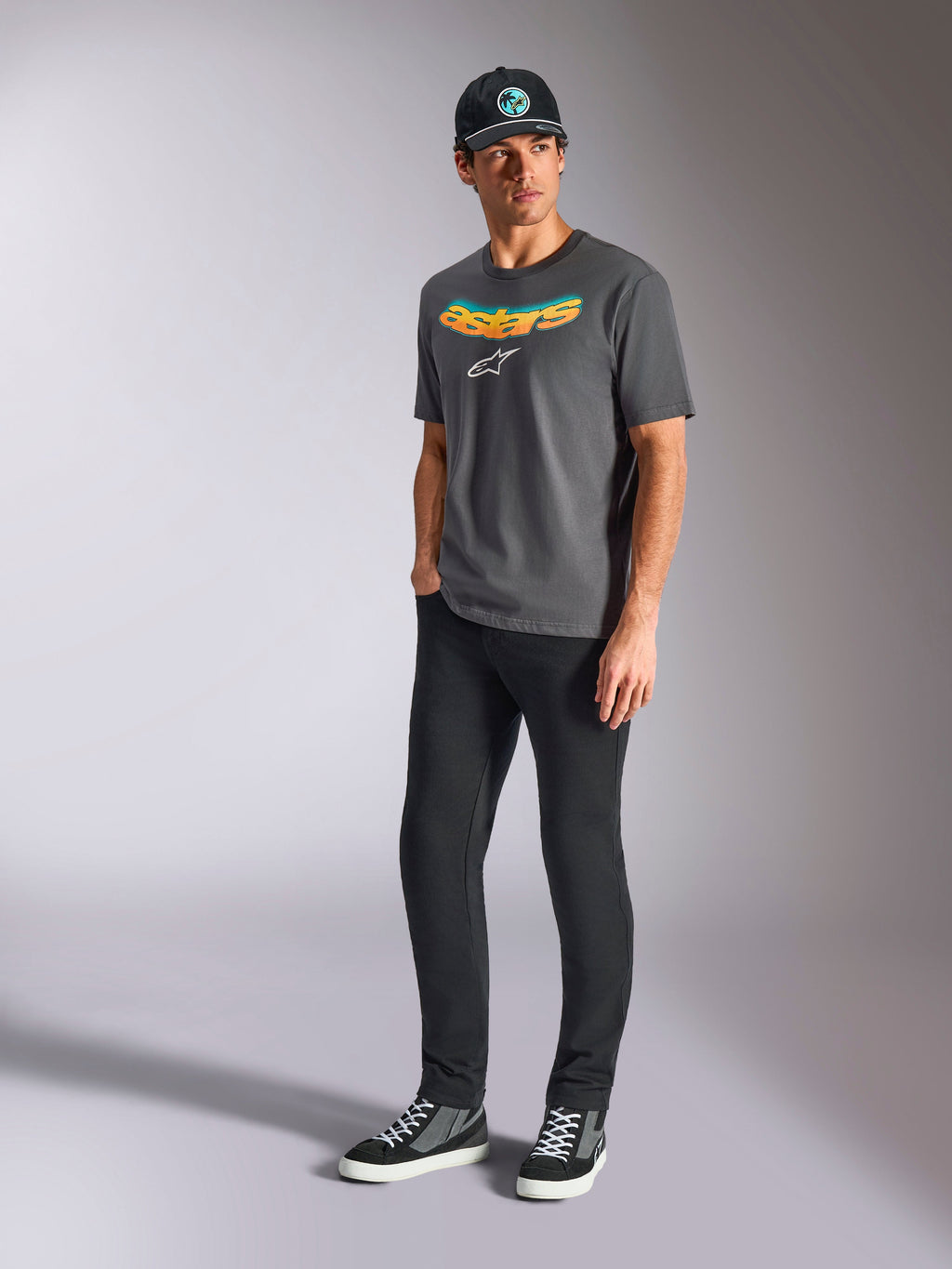 Alpinestars Ellipsoid CSF Tee, t-shirt a maniche corte, grigio antracite, con scritta Astars curva con gradiente di colore e logo bianco, indossata da un modello con pantaloni neri e cappellino, streetwear casual motorsport