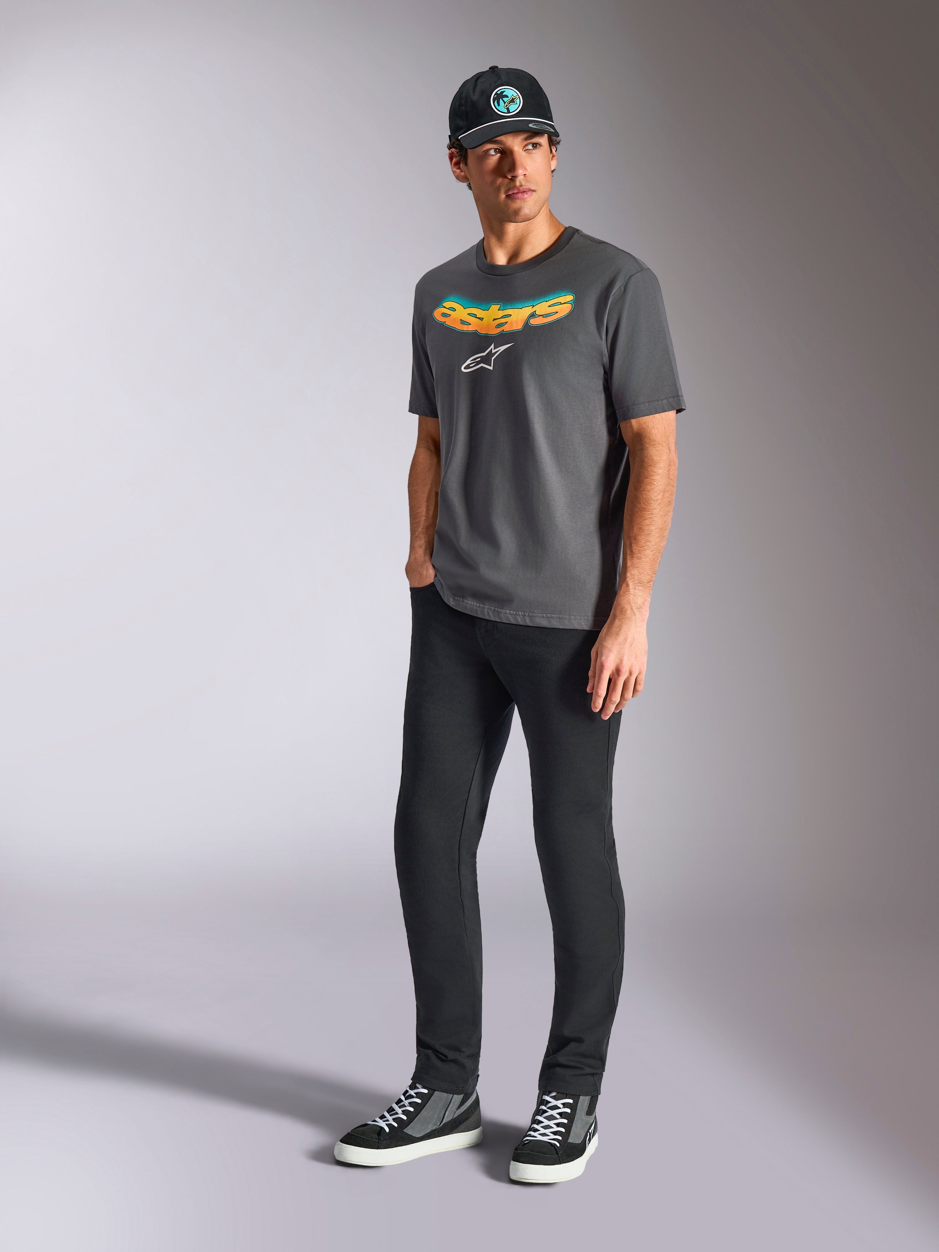 Alpinestars Ellipsoid CSF Tee, t-shirt a maniche corte, grigio antracite, con scritta Astars curva con gradiente di colore e logo bianco, indossata da un modello con pantaloni neri e cappellino, streetwear casual motorsport