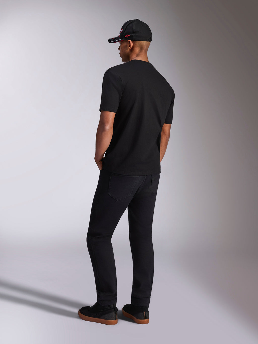 Alpinestars Plate CSF Tee, T-shirt a maniche corte, nera, vista posteriore su modello con pantaloni neri e cappellino da baseball, streetwear casual motorsport