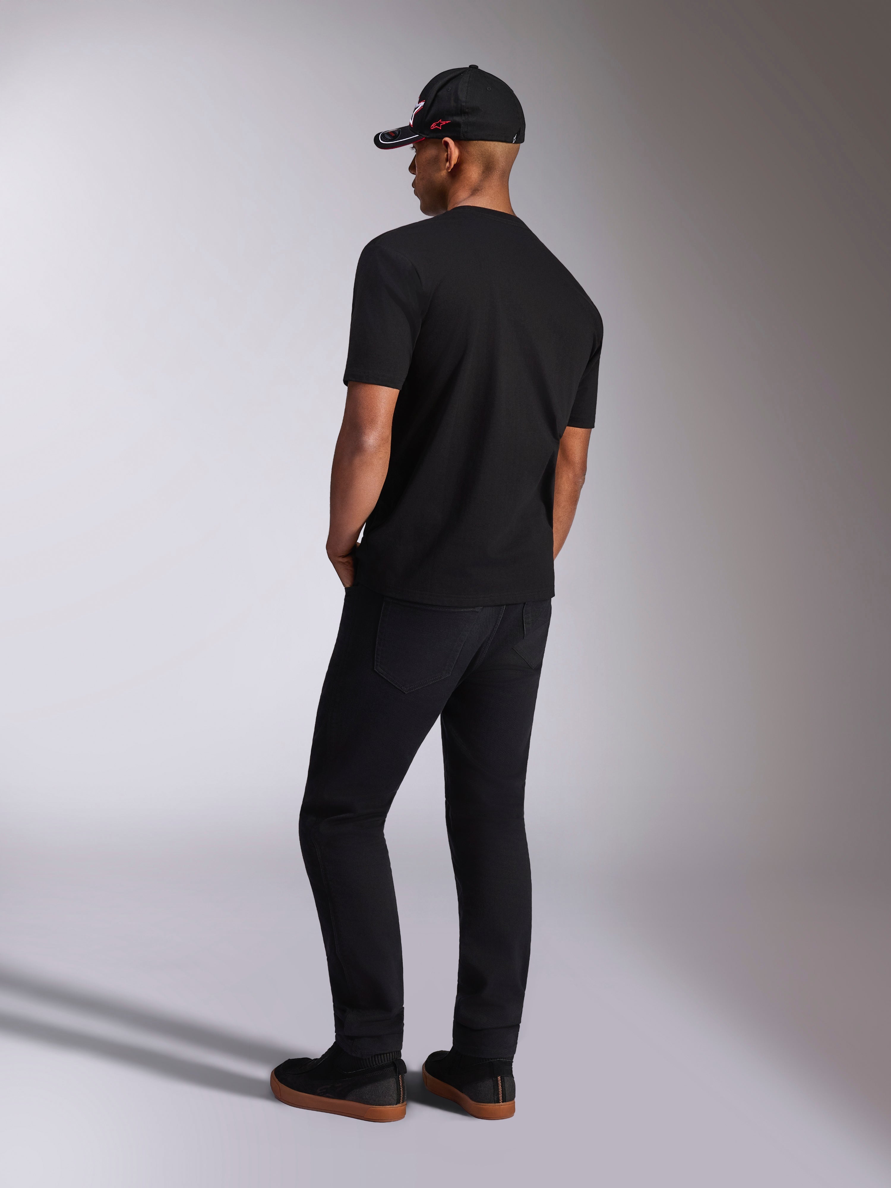 Alpinestars Plate CSF Tee, T-shirt a maniche corte, nera, vista posteriore su modello con pantaloni neri e cappellino da baseball, streetwear casual motorsport
