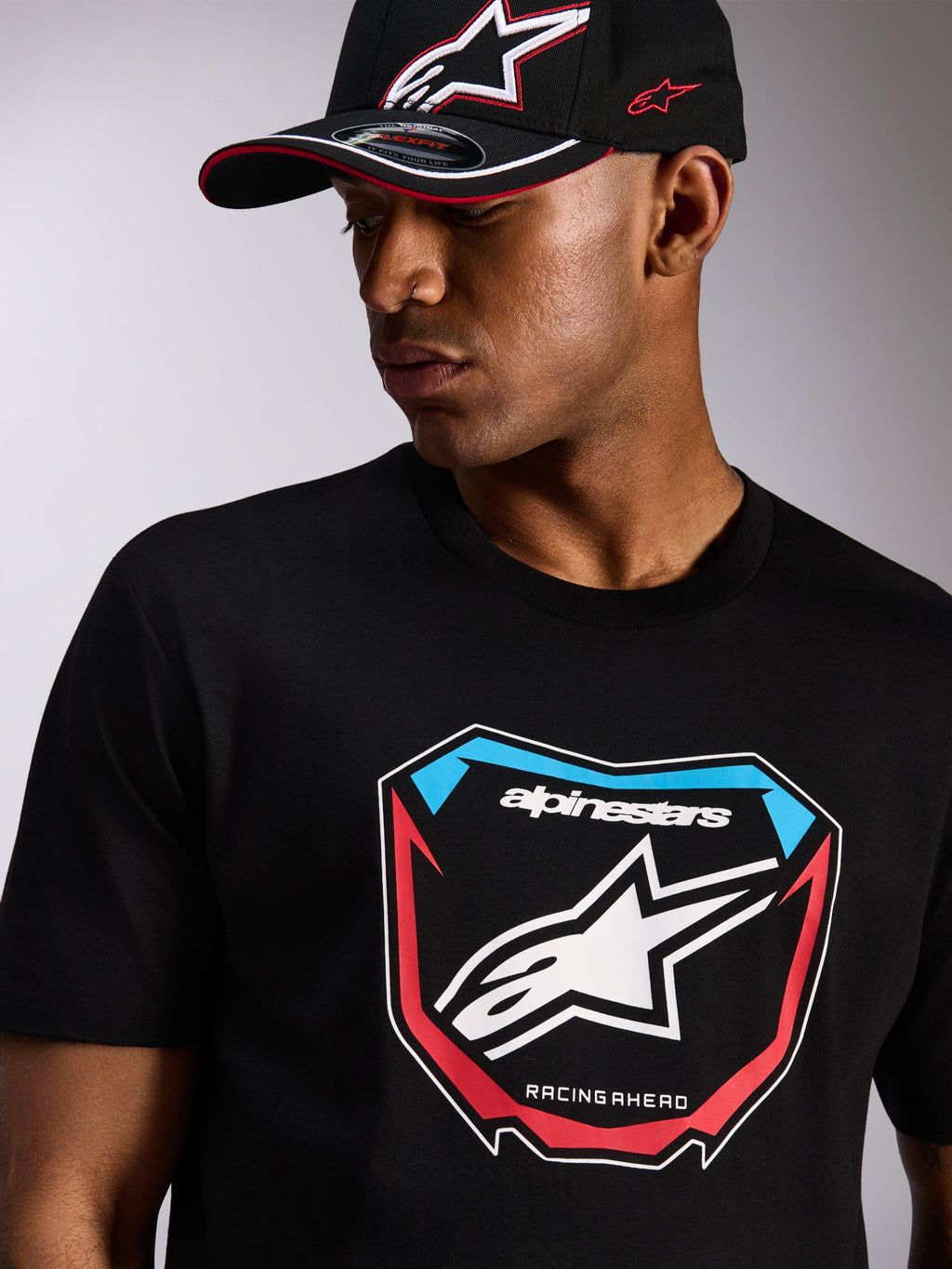 T-shirt Alpinestars Plate CSF, maglietta a maniche corte, nera, grande grafica frontale MX Plate con logo a stella e dettagli rossi e blu, indossata con cappellino Alpinestars nero coordinato, streetwear casual motorsport